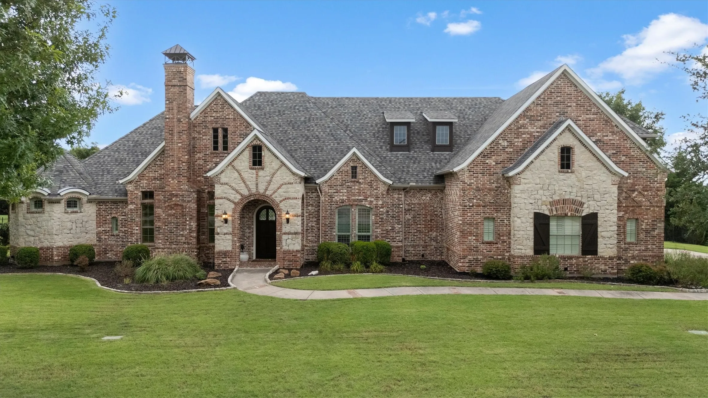 Brick and stone exterior featuring an arched entry, prominent chimney, dormer windows, and dark shutters