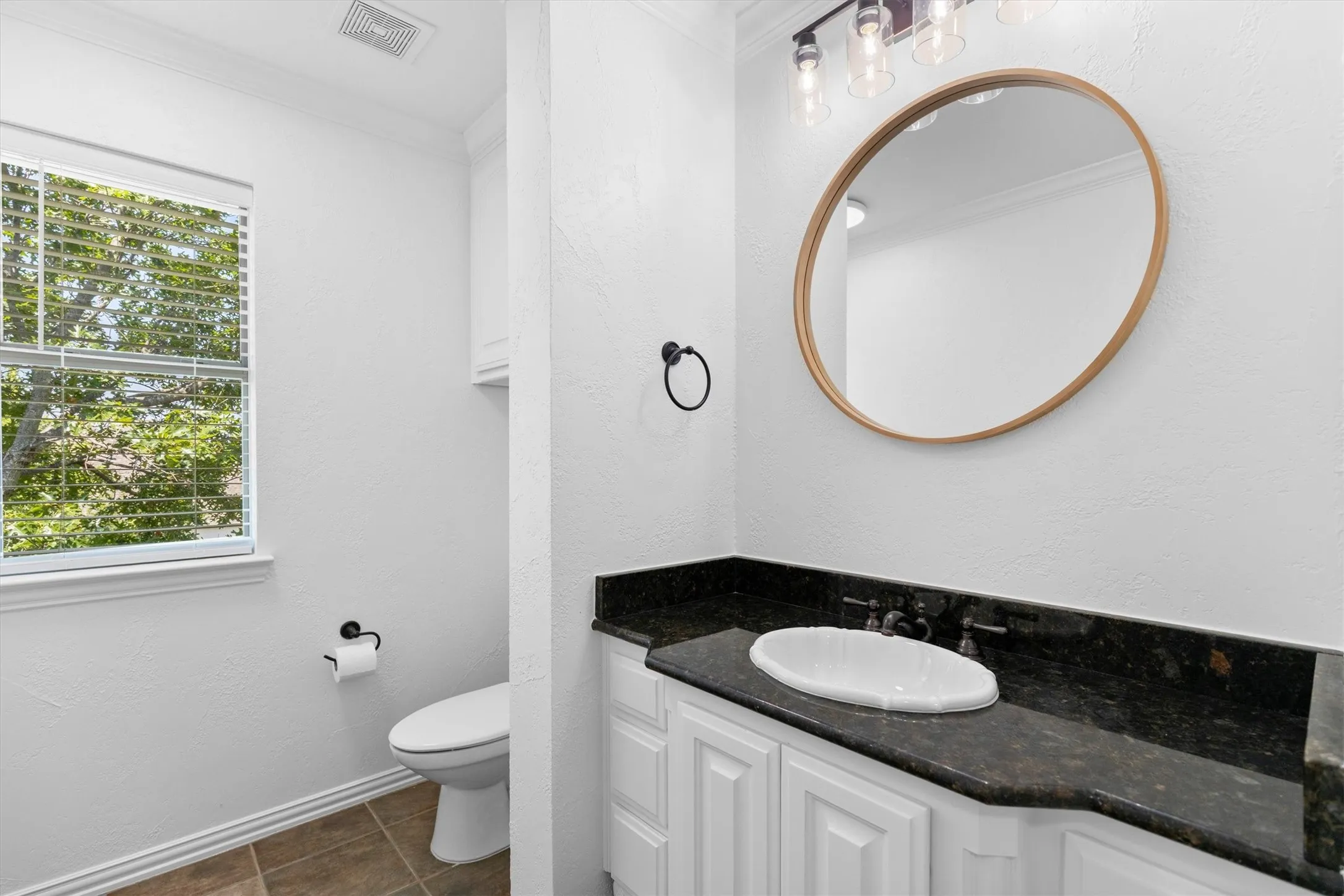 Bathroom featuring a dark granite-style countertop, white cabinetry, and a circular mirror with a wood-finish frame
