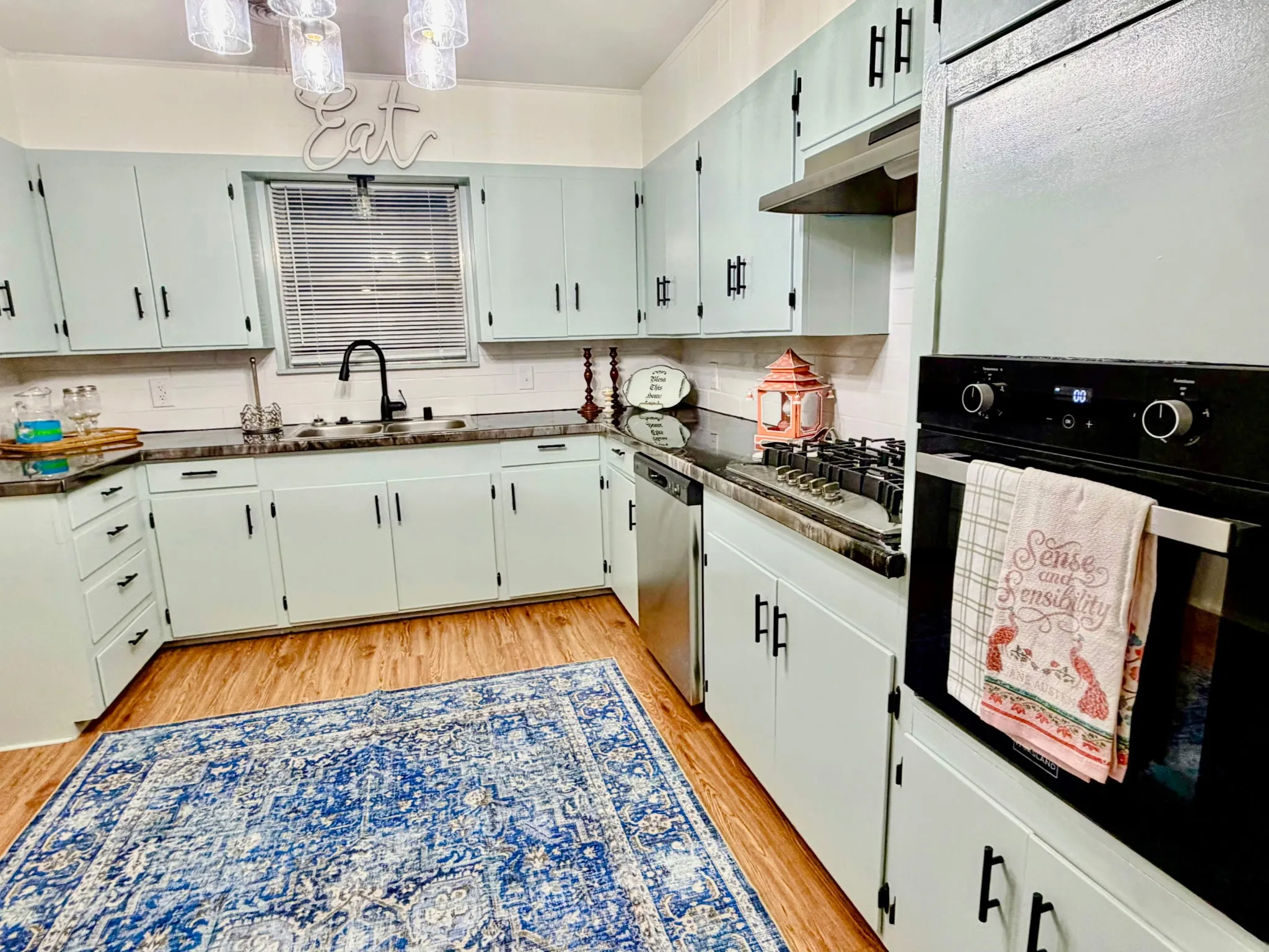 Light mint cabinetry, dark countertops, white tile backsplash, black built-in oven, and wood-finish flooring