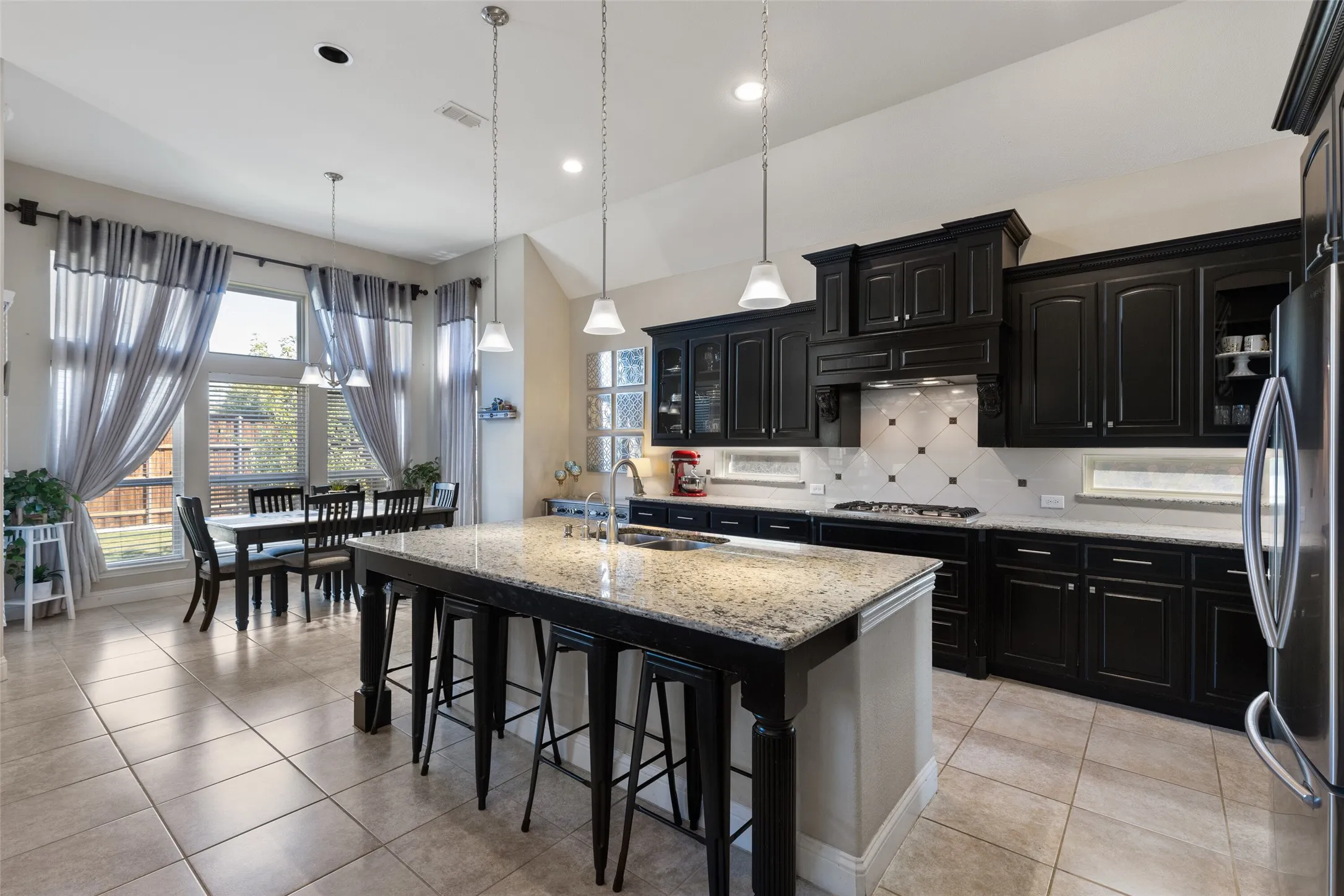 Kitchen featuring dark cabinetry with light stone countertops, a large center island with pendant lighting, a tile backsplash, and stainless steel appliances