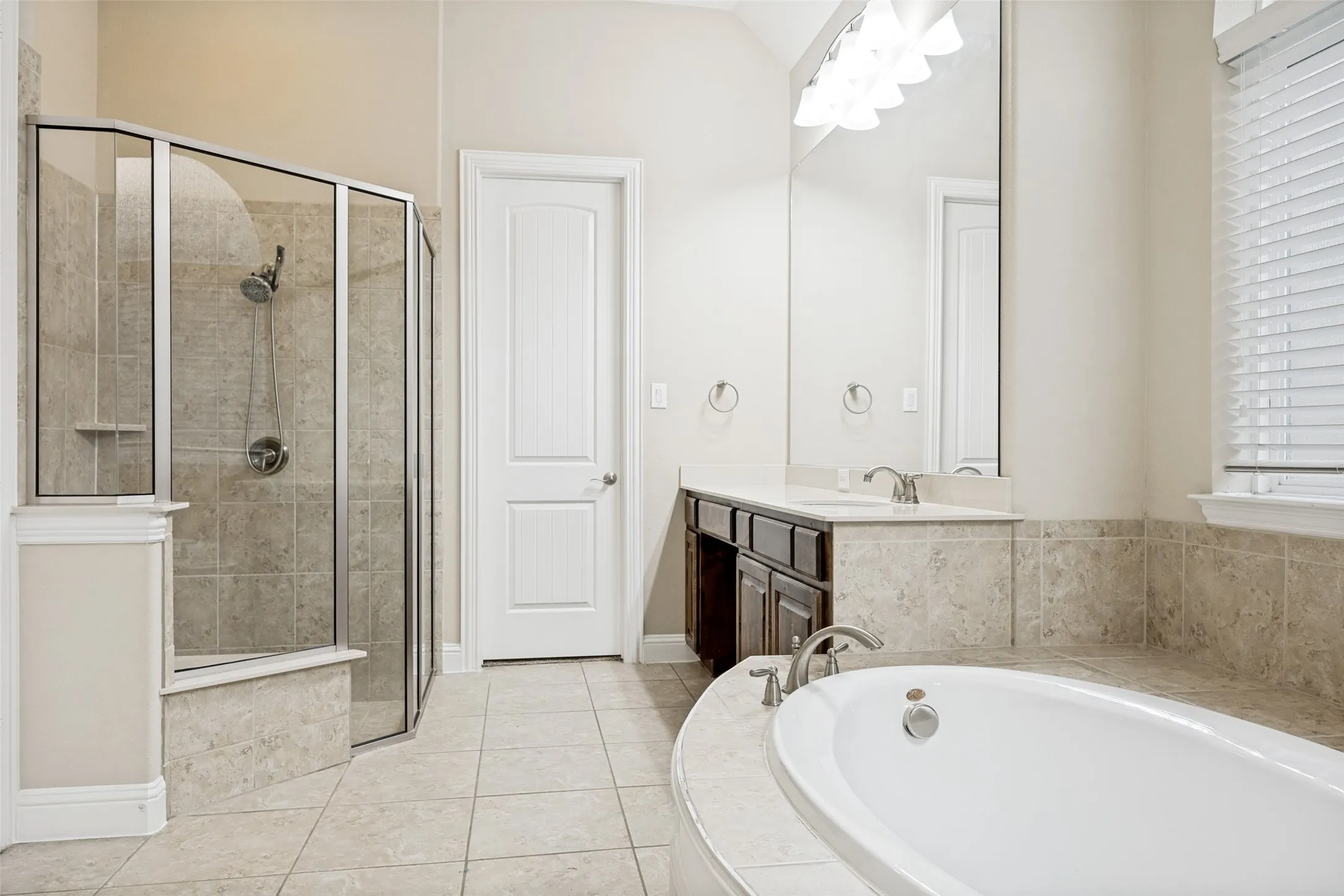 Bathroom featuring an oval soaking tub with deck-mounted faucet, a glass-enclosed shower with tiled walls, a double vanity with two sinks and a large mirror, and tile​​‌​​​​‌​​‌‌​‌‌​​​‌‌​‌​‌​‌​​​‌​​ flooring