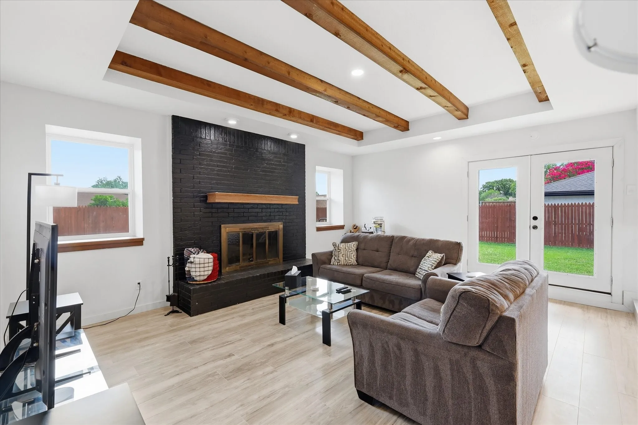 Living area featuring exposed wood ceiling beams, a black brick fireplace with a wood mantle, light wood-finish flooring, and French doors opening to the exterior