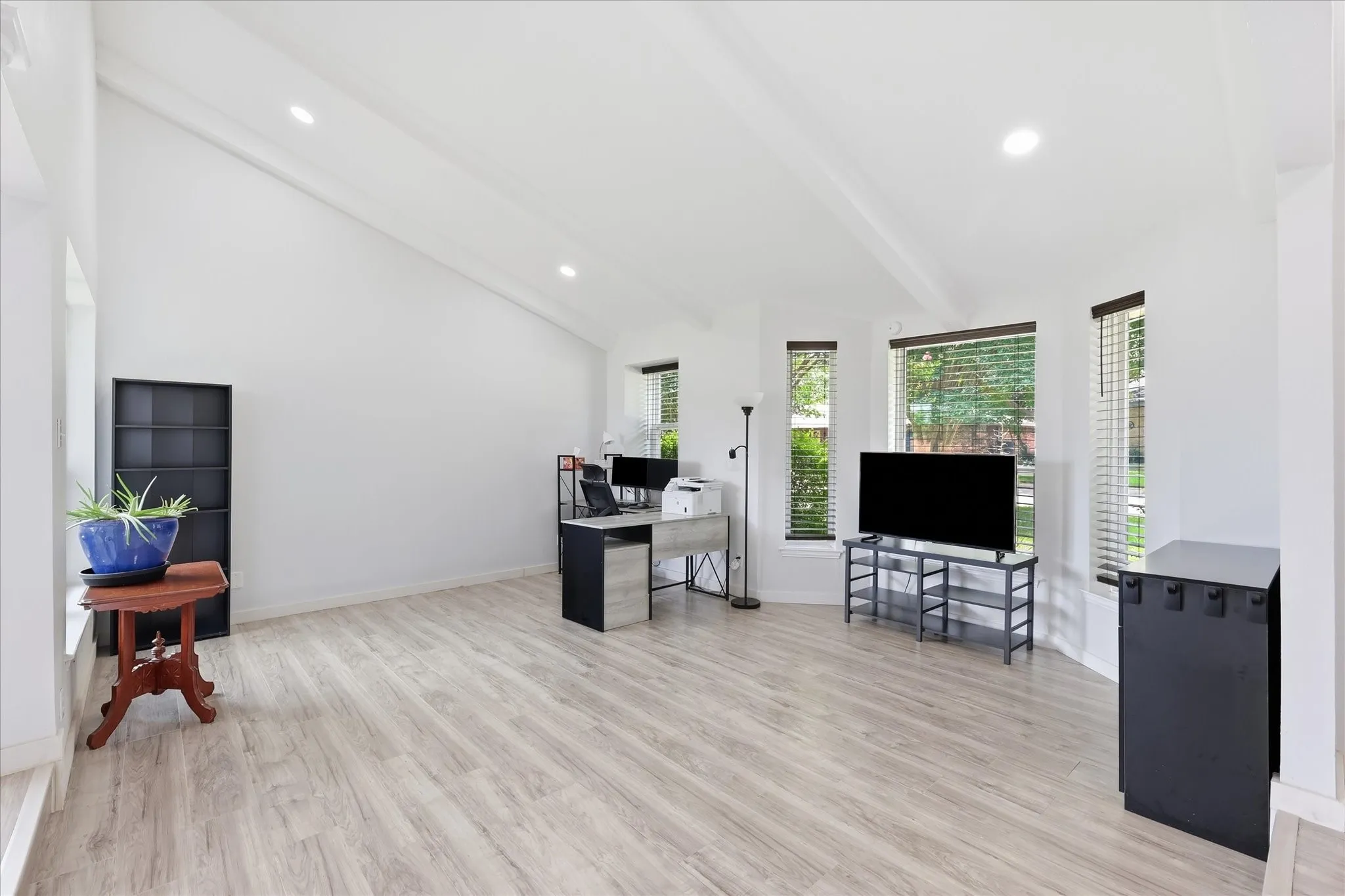 Spacious room featuring vaulted ceilings, recessed lighting, wood-finish flooring, and multiple large windows with horizontal blinds