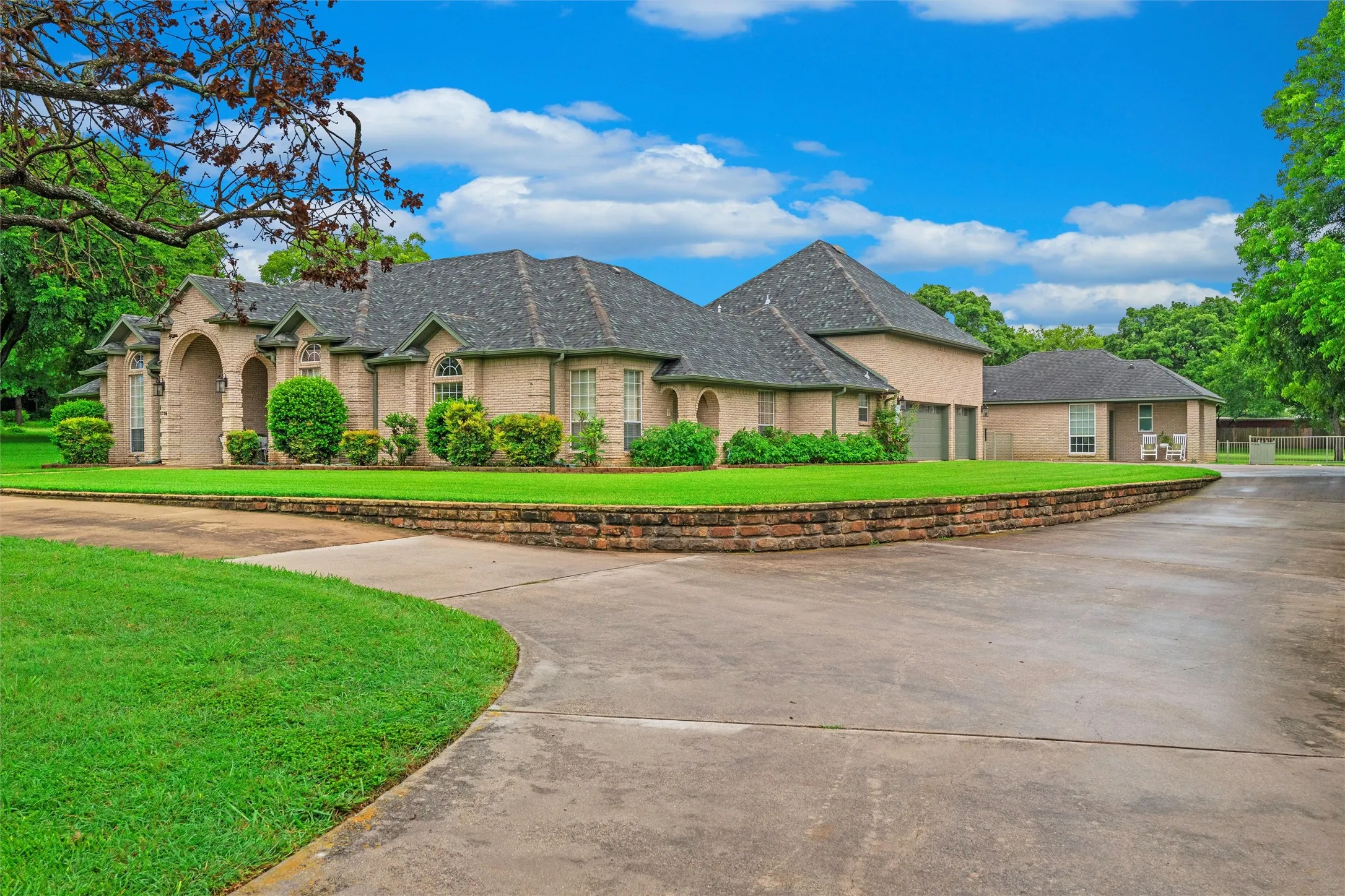 Expansive front driveway featuring a light-colored brick facade, a shingled roof, arched entryway, and multiple garage doors