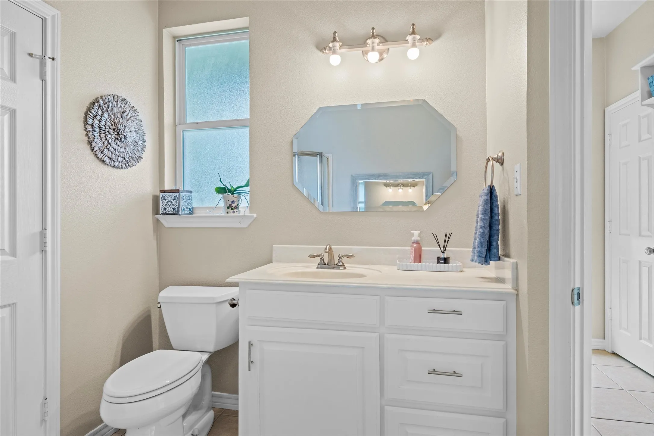 Bathroom featuring a white vanity with a light-colored countertop, an octagonal mirror, a three-light vanity fixture, a toilet, and a window with frosted glass