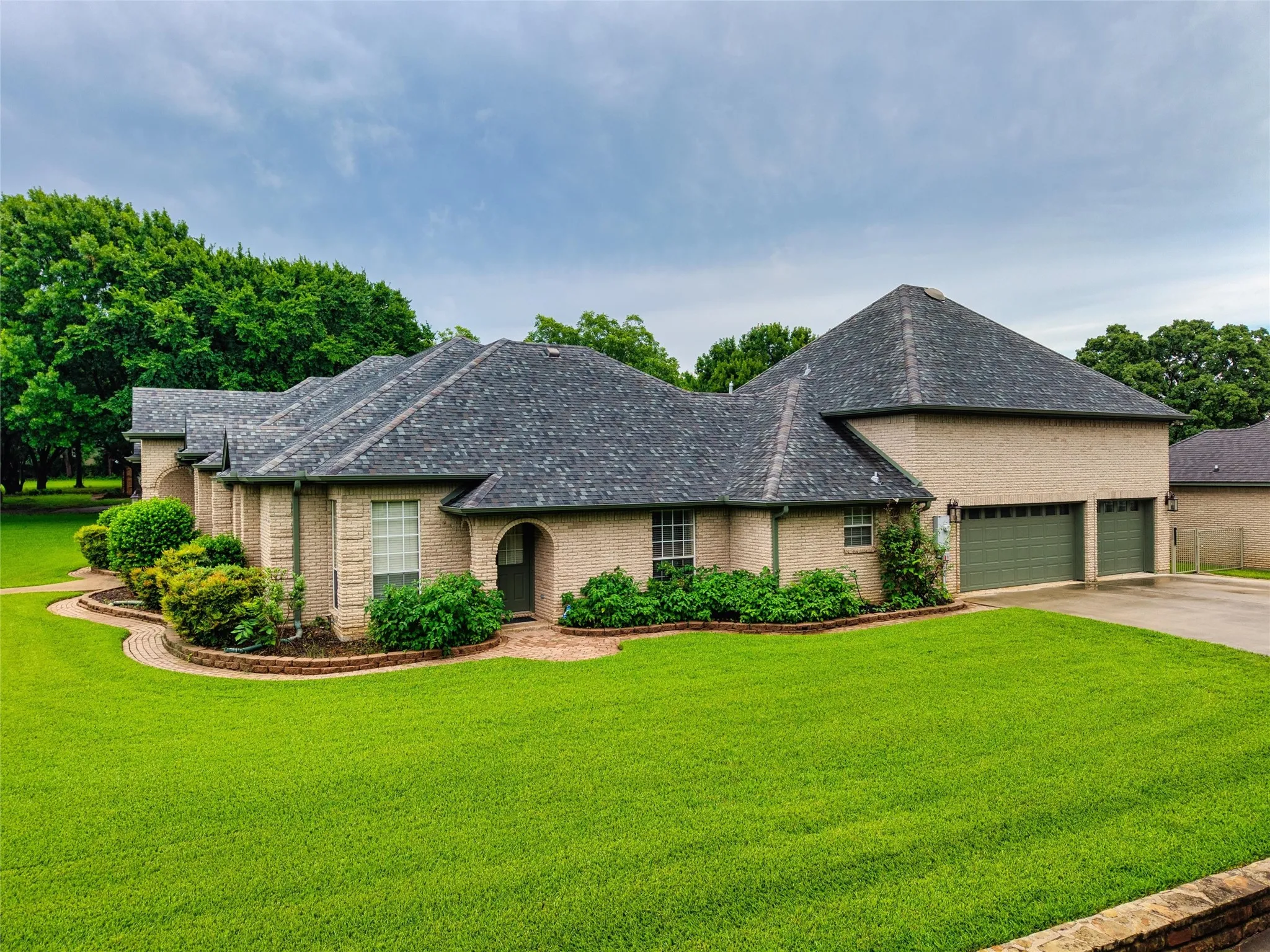 Spacious brick exterior home featuring a lush, well-maintained lawn, established landscaping, arched entryway, and an attached multi car garage with matching green doors