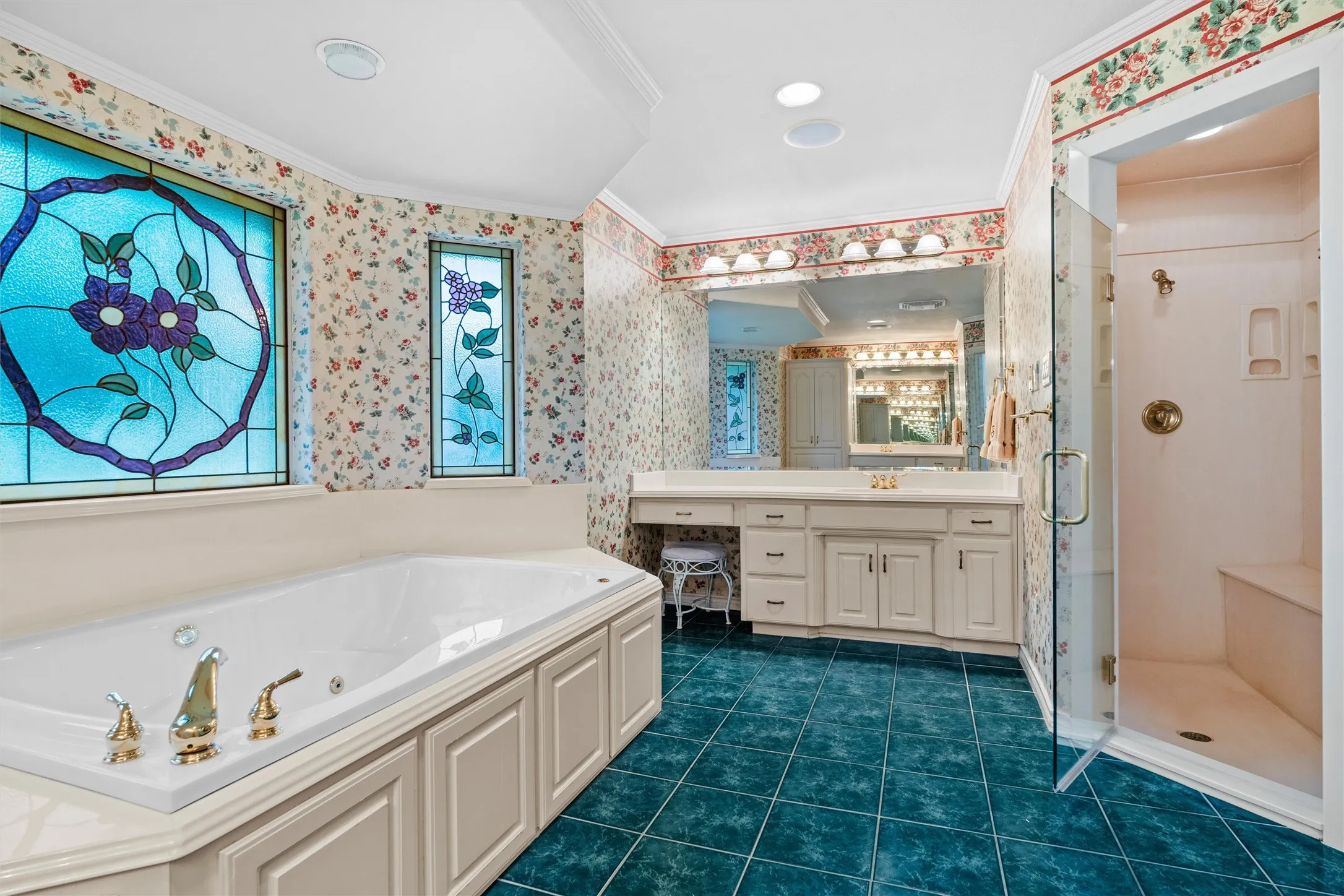 Spacious bathroom featuring a built-in jetted tub with brass fixtures, stained glass windows, a glass-enclosed shower with bench seating, and a double vanity with ample storage,.