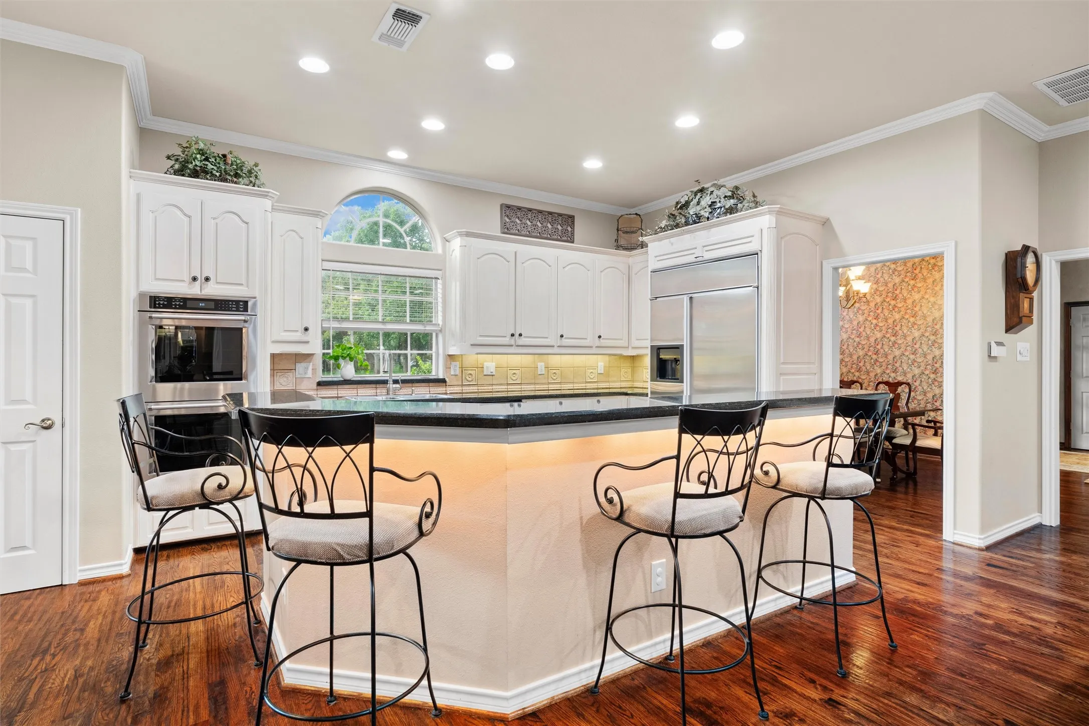 Kitchen featuring expansive island, double oven, white cabinetry, granite countertops, built-in stainless steel appliances, wood-finish flooring, and a large arched window