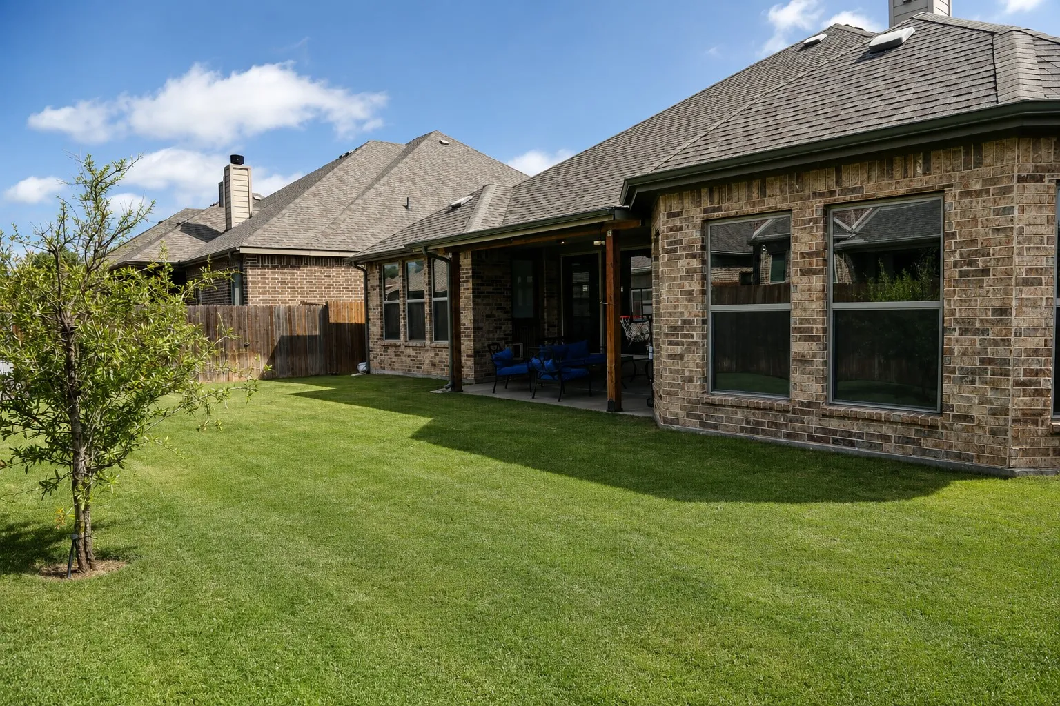 Expansive green lawn featuring a young tree, brick exterior, covered patio area, and large windows