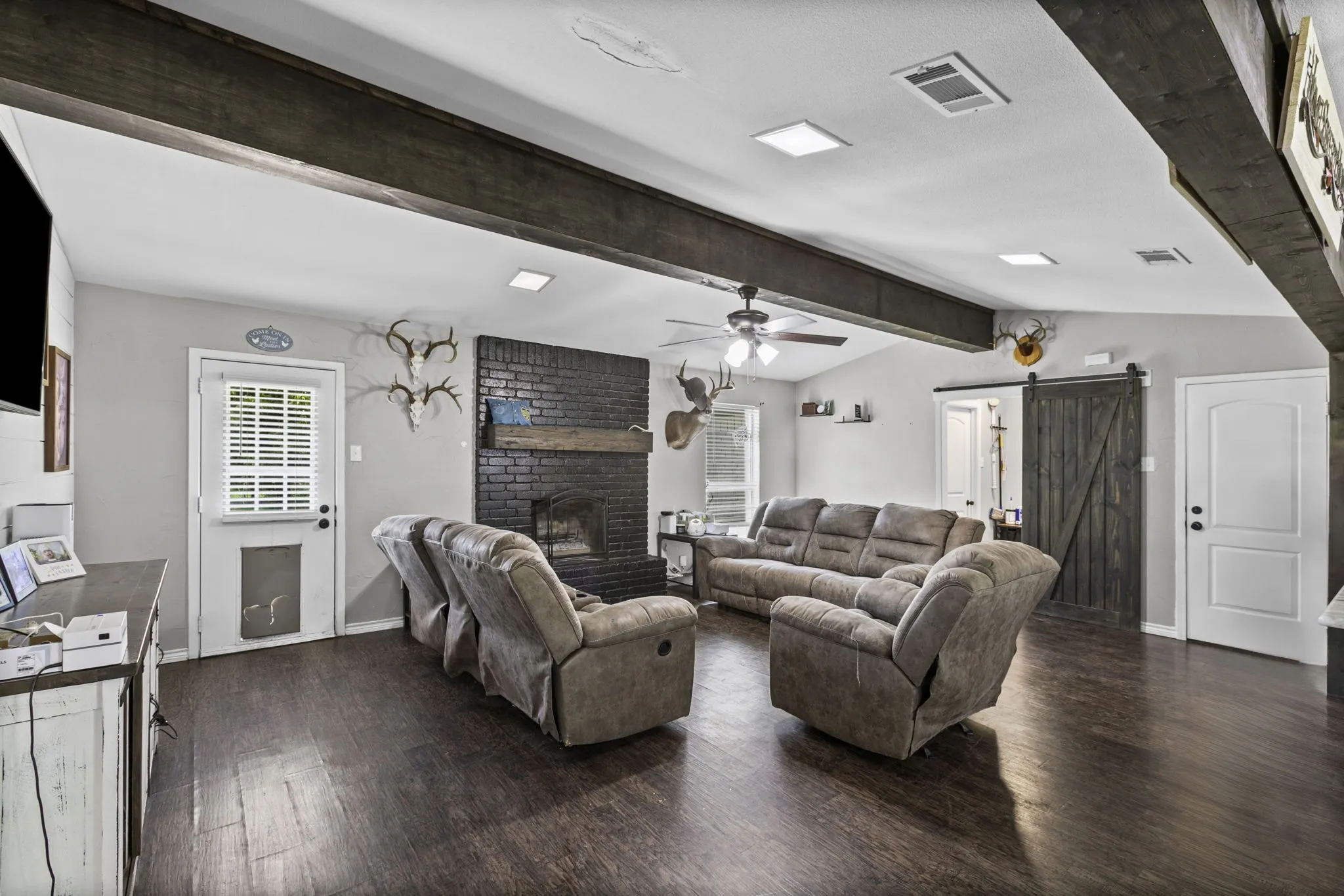Spacious living area featuring exposed ceiling beams, dark wood-finish flooring, and a black brick fireplace with a wood mantle