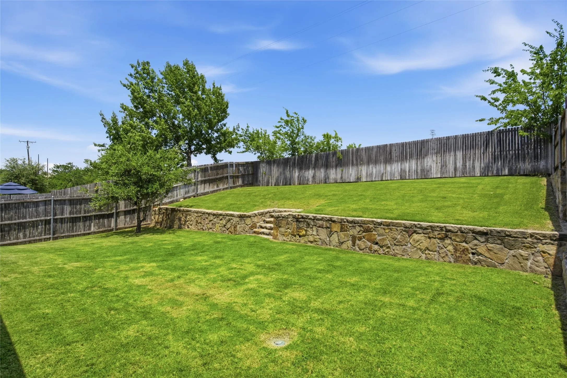 Expansive terraced backyard featuring a stone retaining wall, mature trees, and a wooden privacy fence