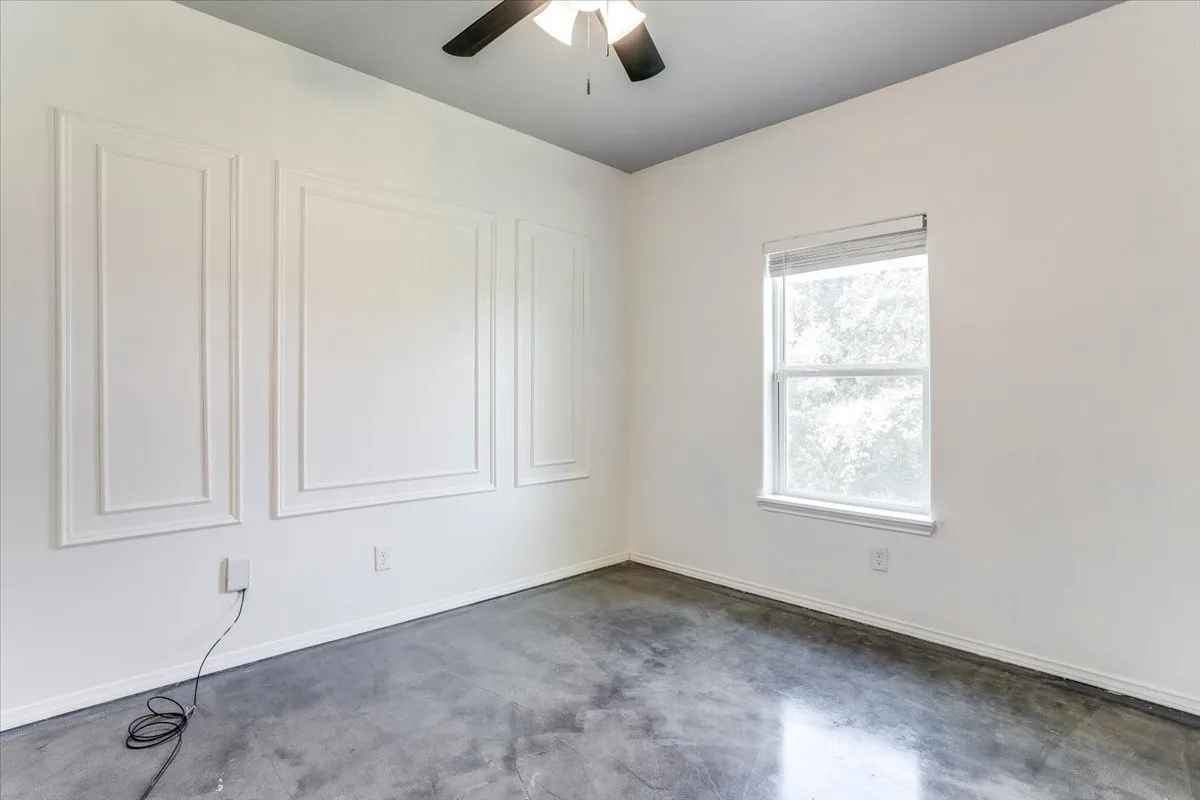 Room featuring accent wall with decorative wall molding, a single window with blinds, dark-toned ceiling fan, and concrete flooring