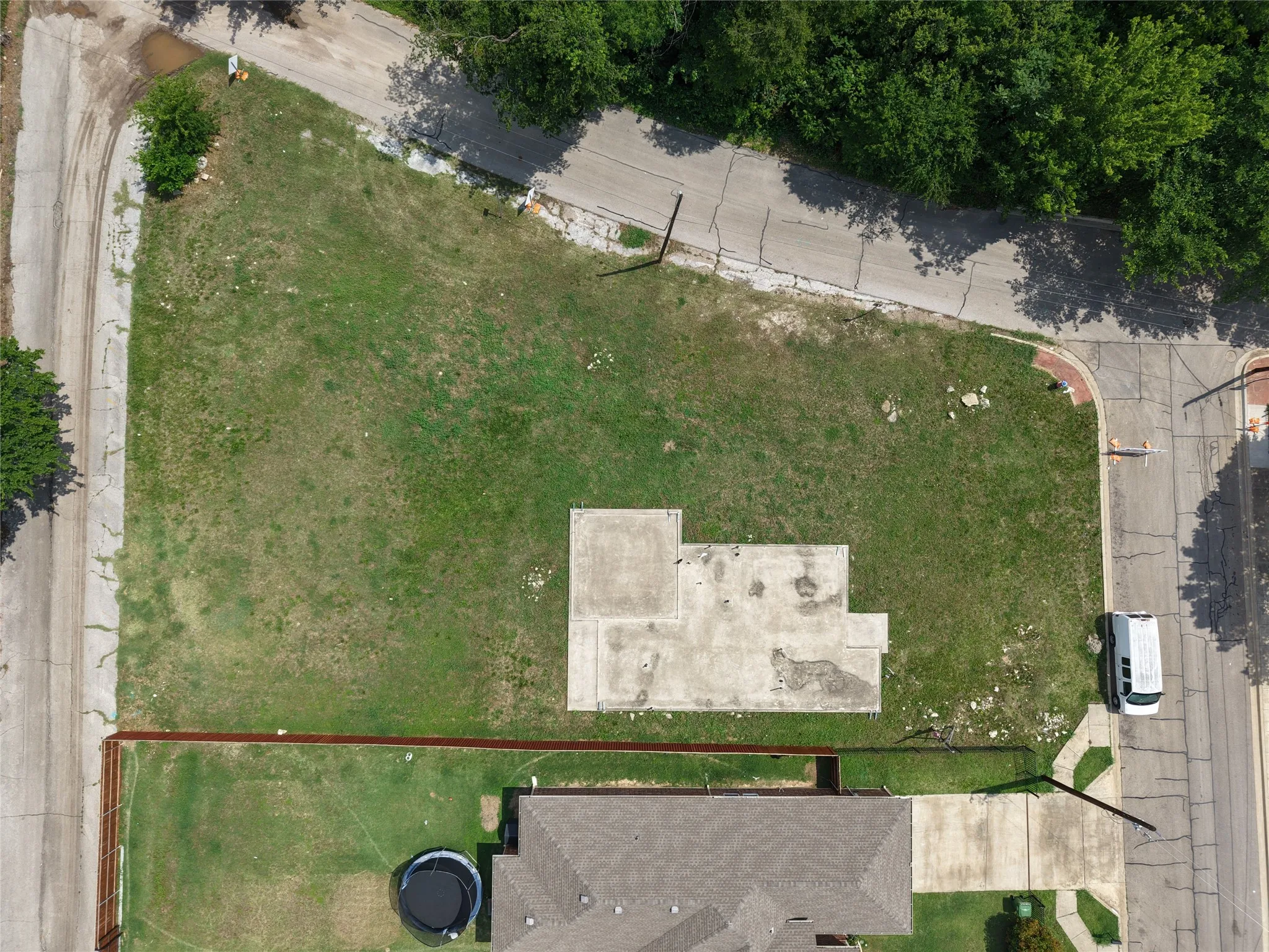 Expansive corner lot featuring a concrete foundation, mature trees bordering one side, and two distinct paved road frontages