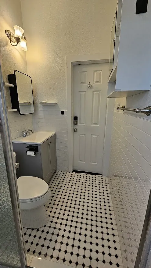 Bathroom featuring black and white mosaic tile flooring, a gray vanity with an integrated sink, a wall-mounted mirror with shelving, subway tile wainscoting, and upper cabinetry