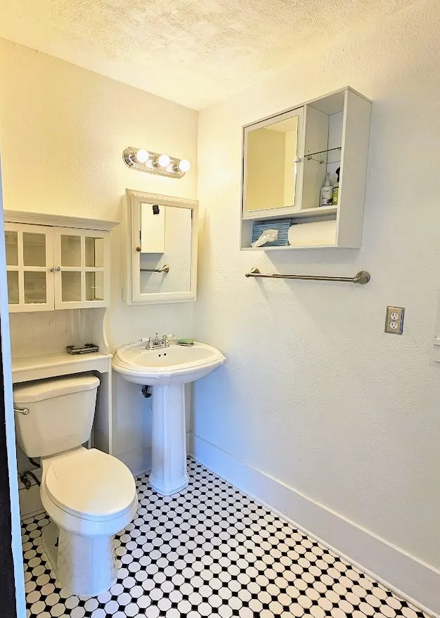 Bathroom featuring black and white hexagonal tile flooring, a pedestal sink with a chrome faucet, a wall-mounted medicine cabinet, and a built-in toilet-side storage unit