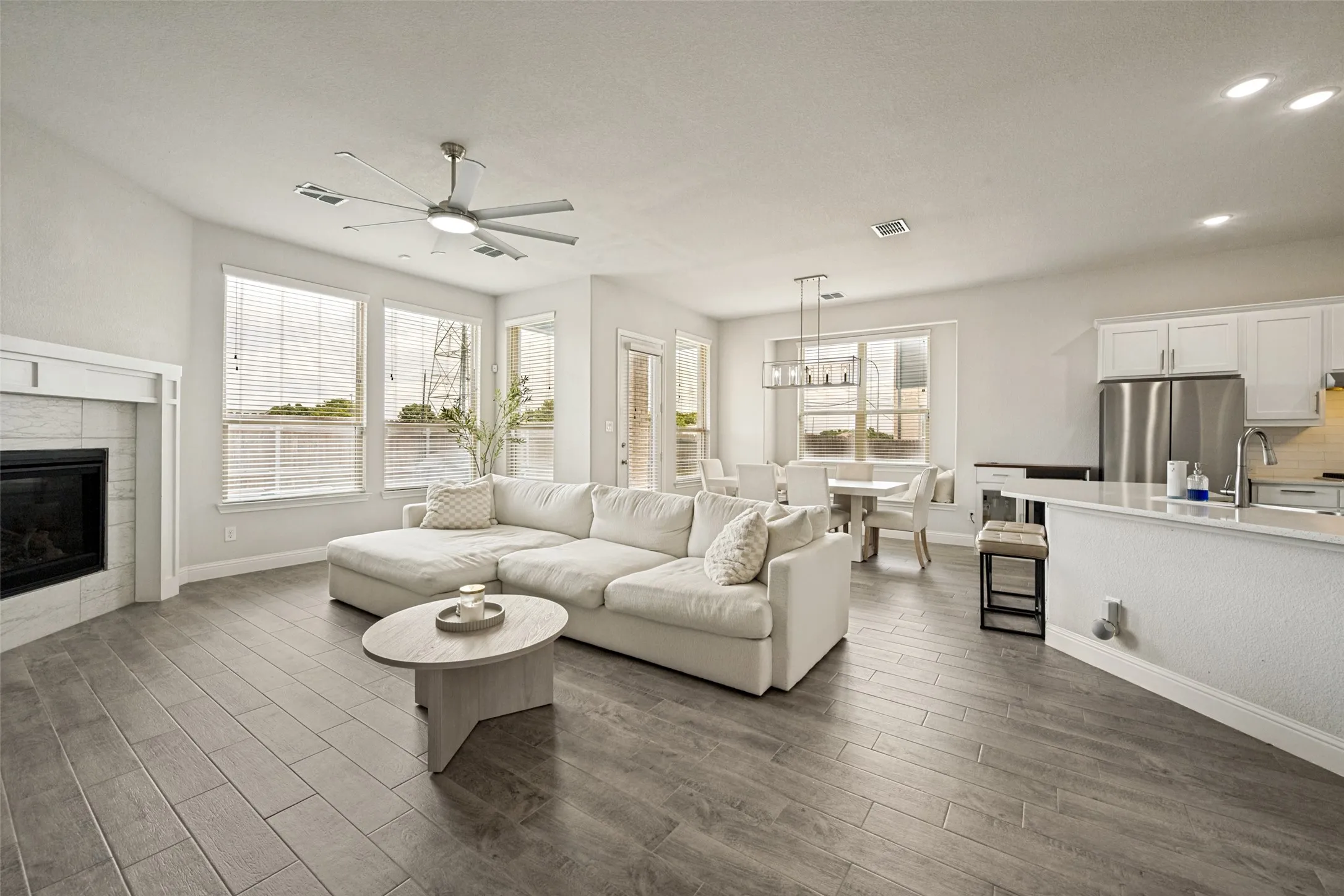 Spacious open-concept living area featuring wood-finish flooring, a contemporary fireplace with mantel, and abundant natural light from multiple windows
