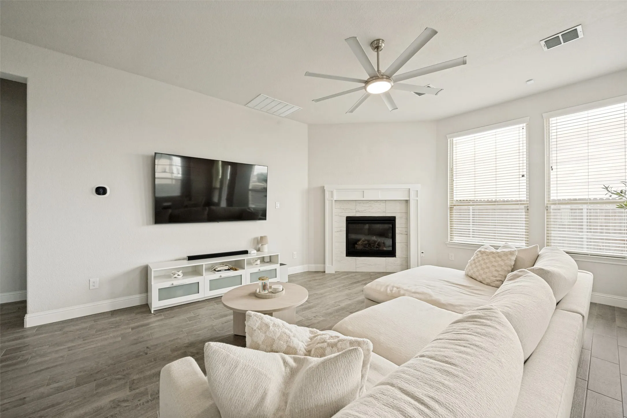 Living area featuring wood-finish flooring, a tiled fireplace with mantel, multiple large windows, and a ceiling fan with integrated lighting