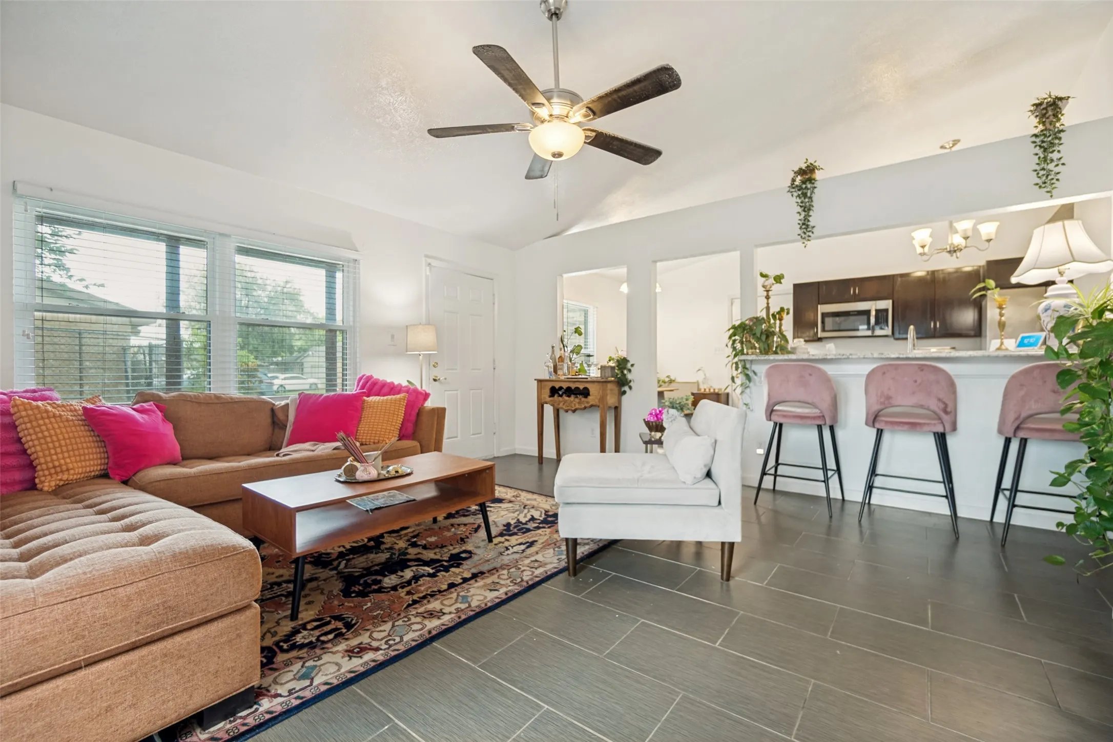 Spacious living area featuring vaulted ceilings, ceiling fan with integrated lighting, large windows with blinds, and dark gray tile flooring