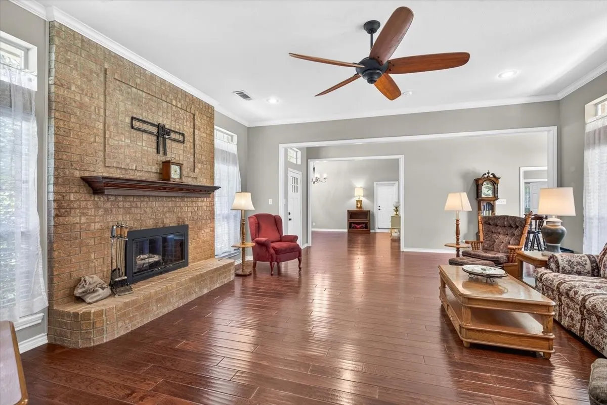 Living area featuring a brick fireplace with a wood mantle, wood-finish flooring, a ceiling fan, and crown molding