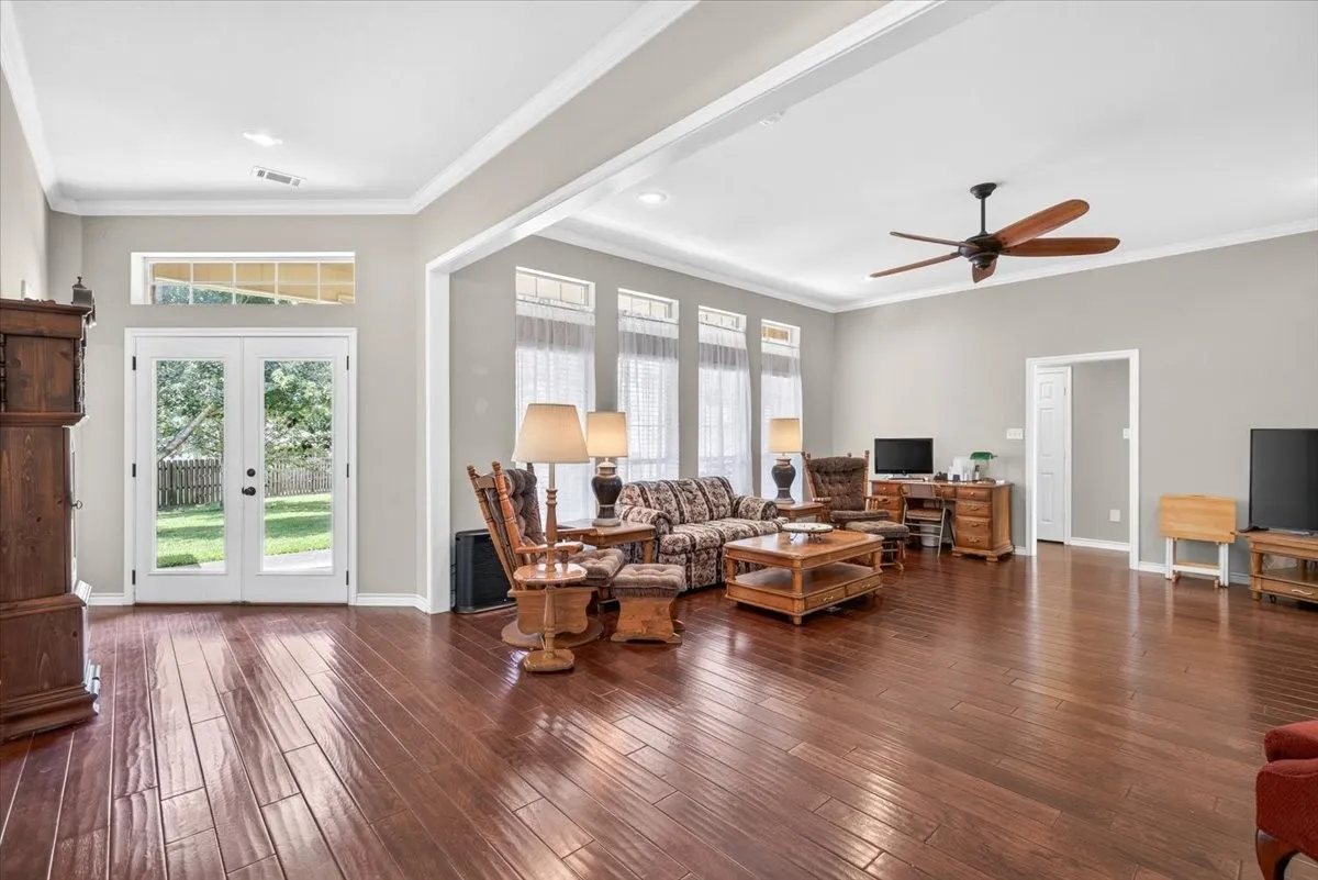 Spacious living area featuring polished wood-finish flooring, a tray ceiling with a ceiling fan, and extensive natural light from multiple​​‌​​​​‌​​‌‌​‌‌​​​‌‌​‌​‌​‌​​​‌​​ windows