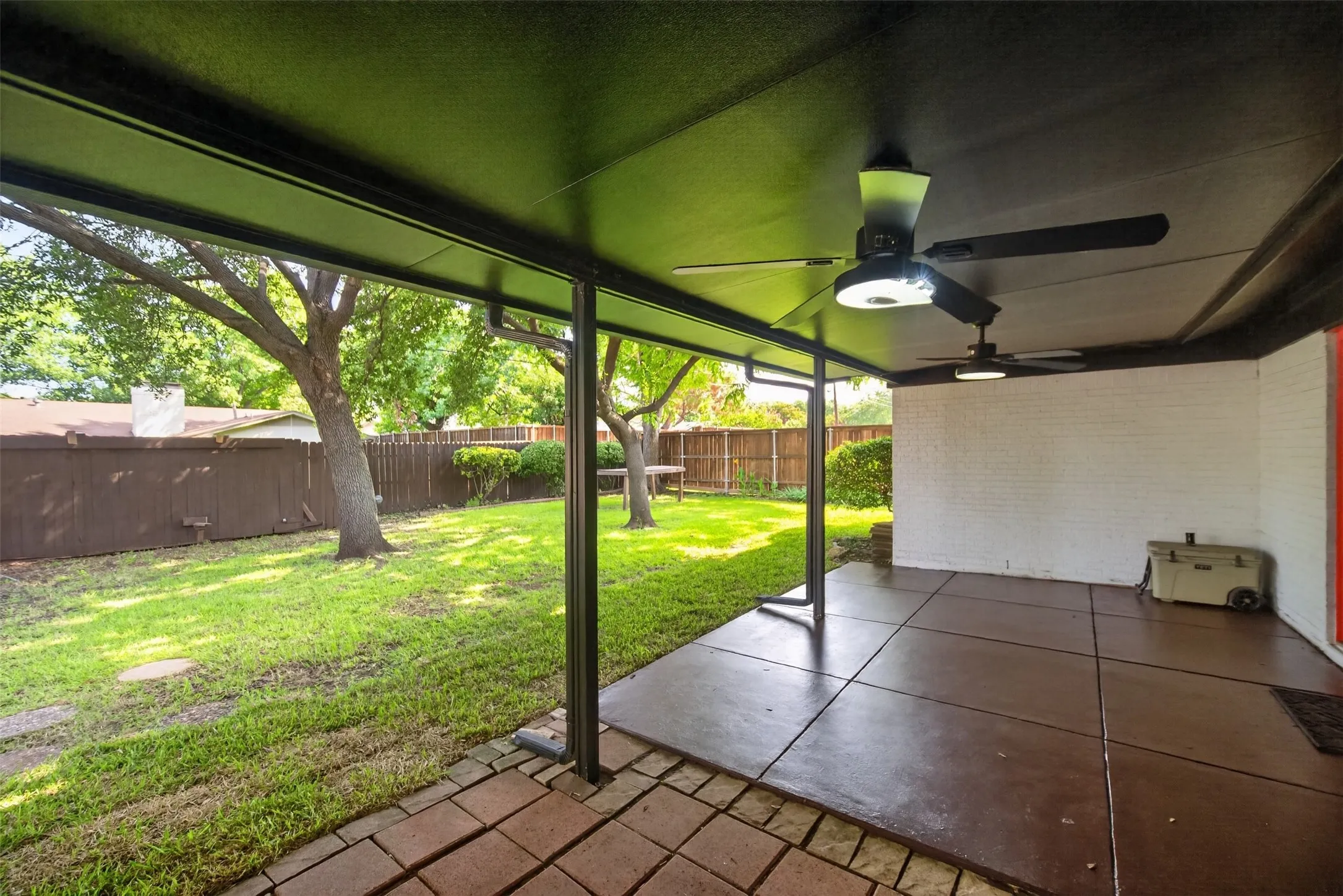 Spacious covered patio featuring two ceiling fans with integrated lighting, a concrete slab, and brick paver flooring