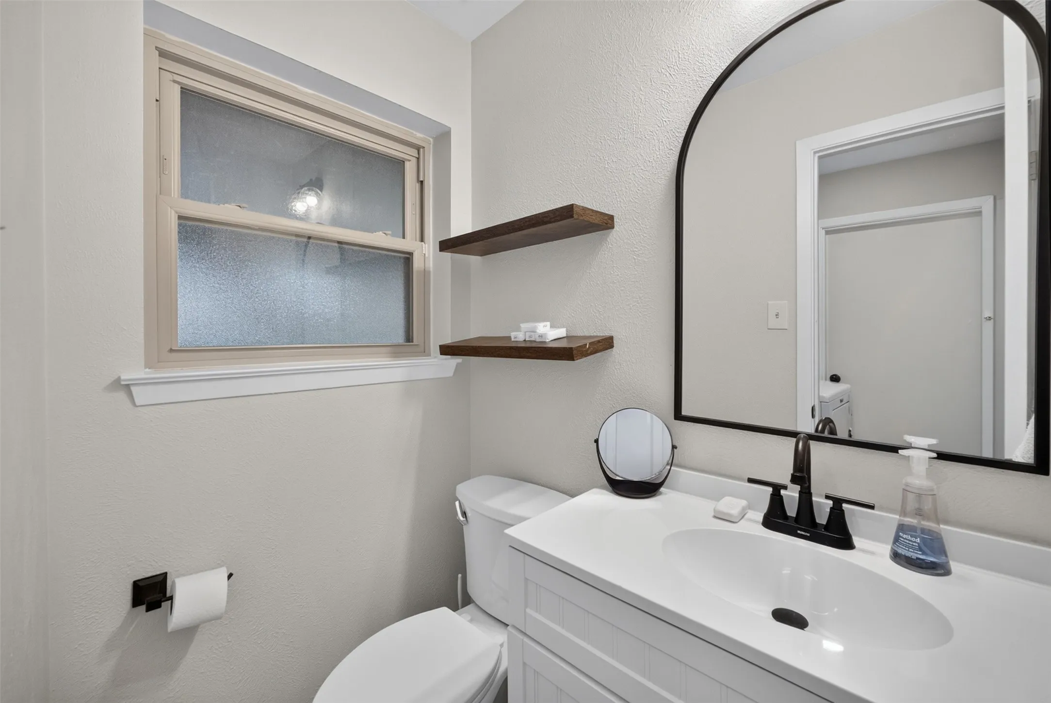Bathroom vanity featuring a white countertop with an integrated oval sink, matte black faucet, and an arched black-framed mirror