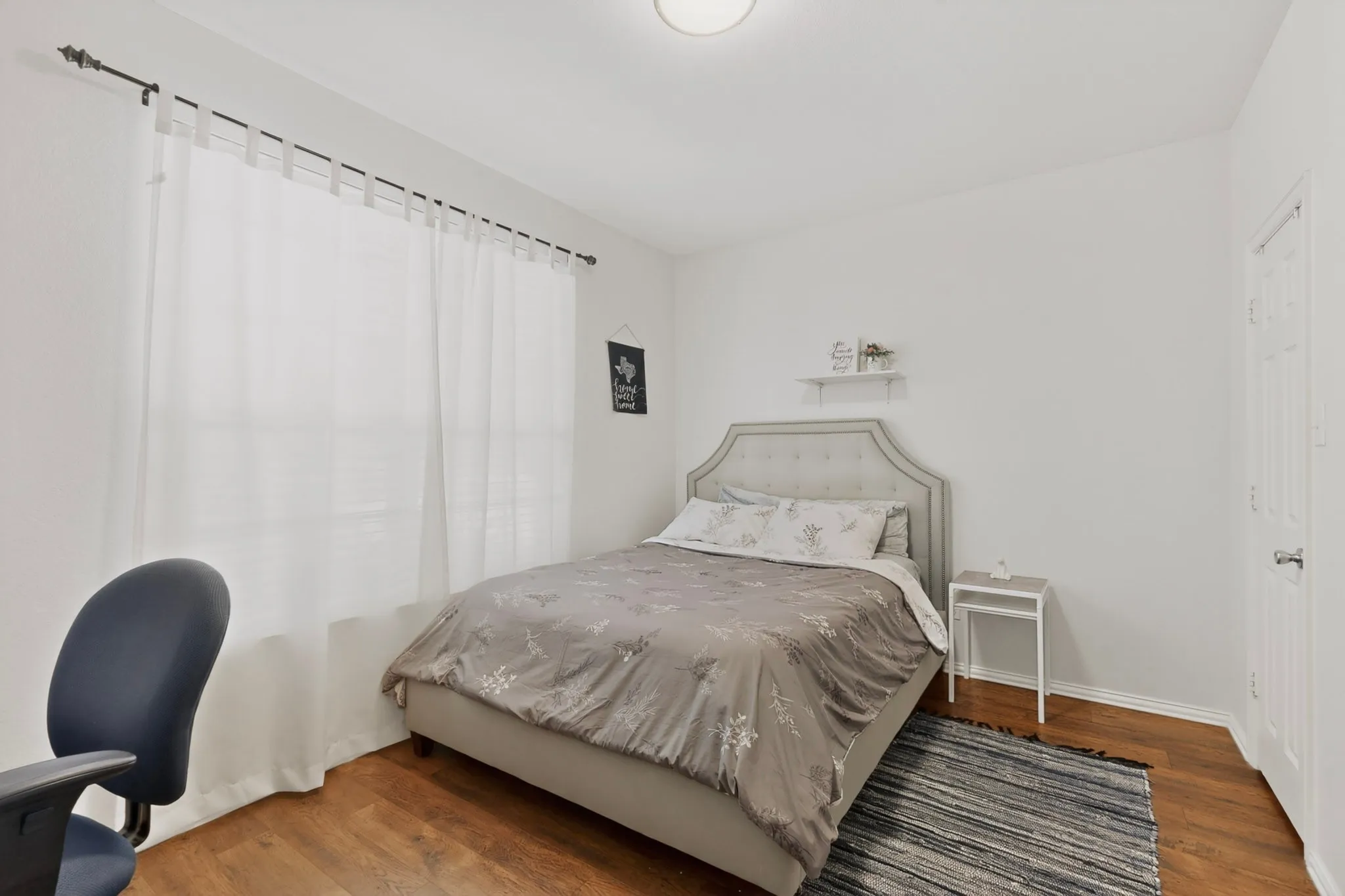Room featuring wood-finish flooring, a contemporary ceiling light fixture, white walls, and a window with sheer white curtains on a dark curtain rod