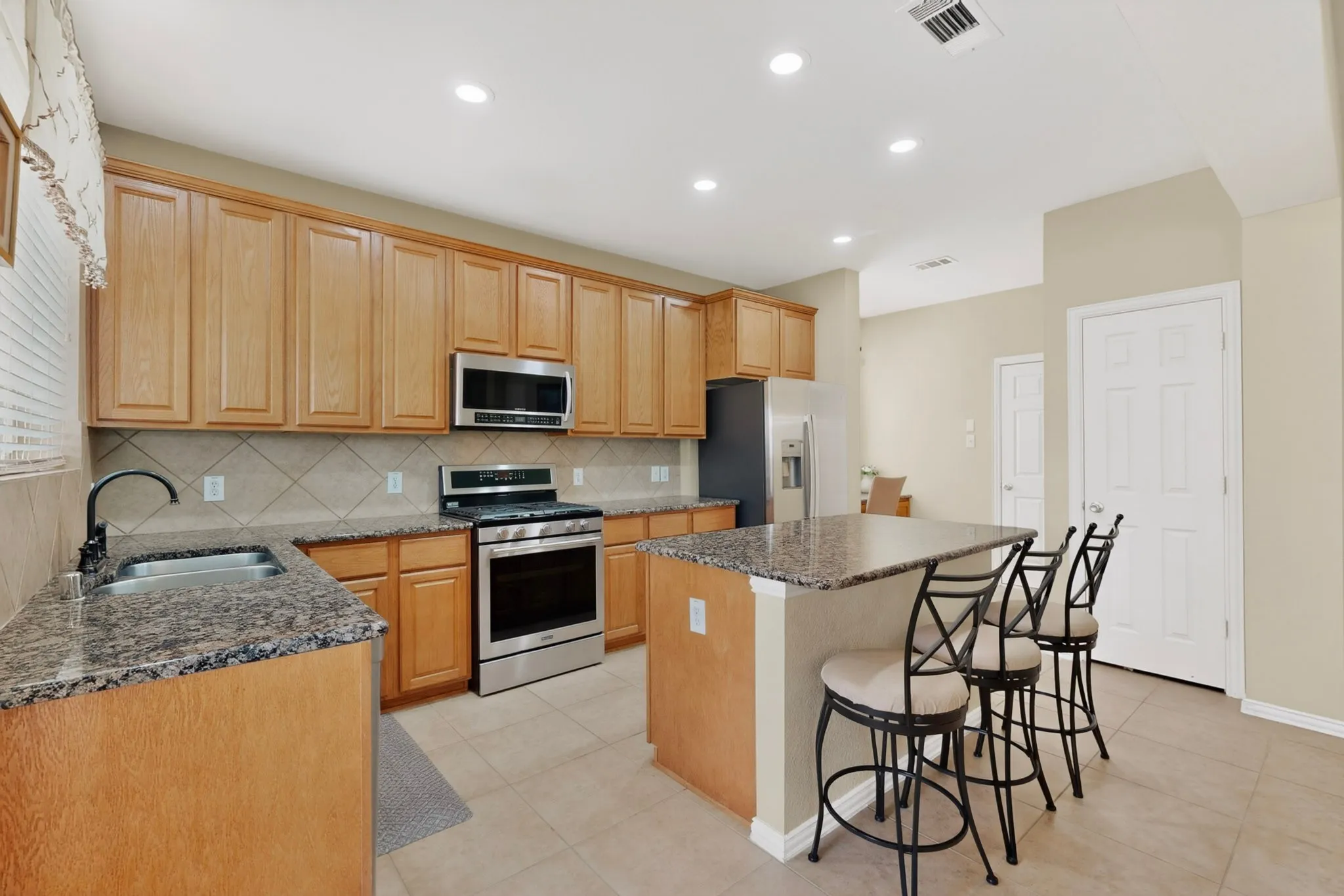 Kitchen featuring light wood cabinetry, granite countertops, tile backsplash, stainless steel appliances, and a central island with seating