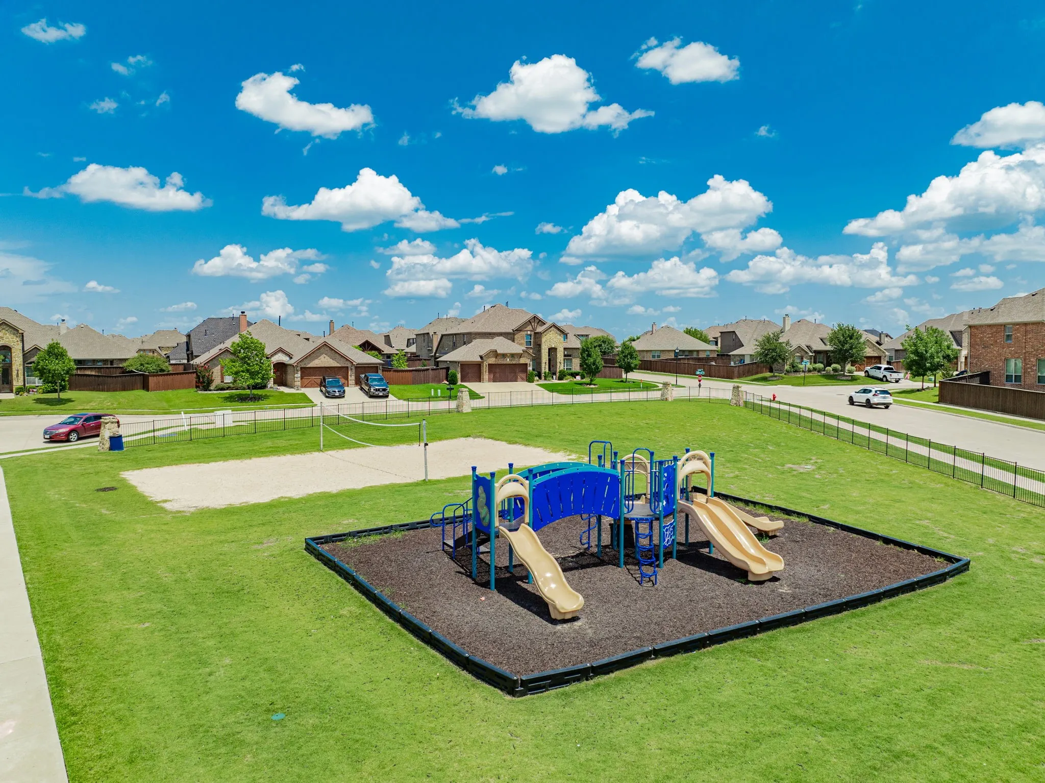 Community greenspace featuring a playground structure with slides and climbing elements on a soft surface, alongside a sand volleyball court