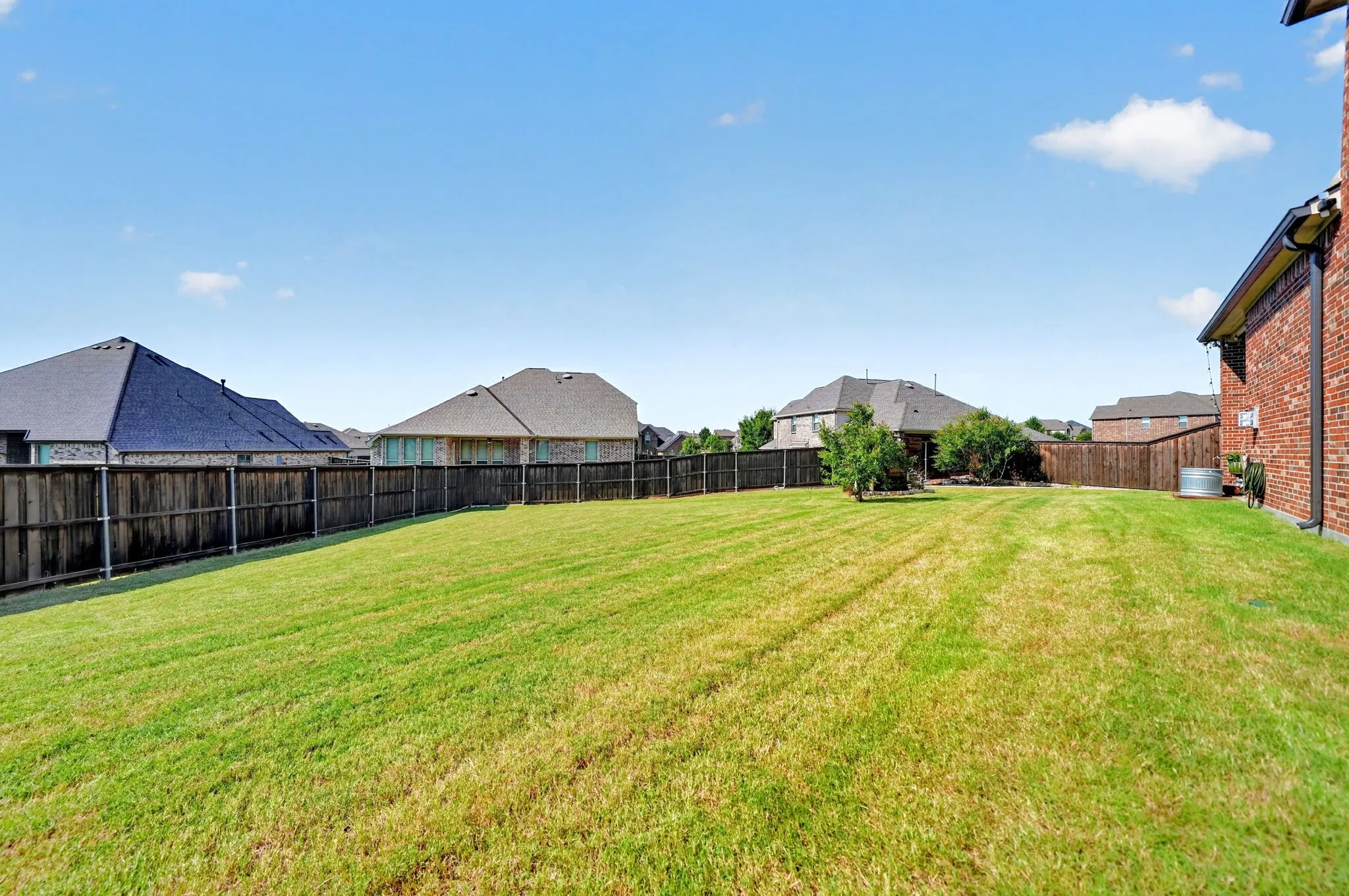 Expansive grassy backyard featuring a full privacy fence, mature landscaping, and a brick exterior