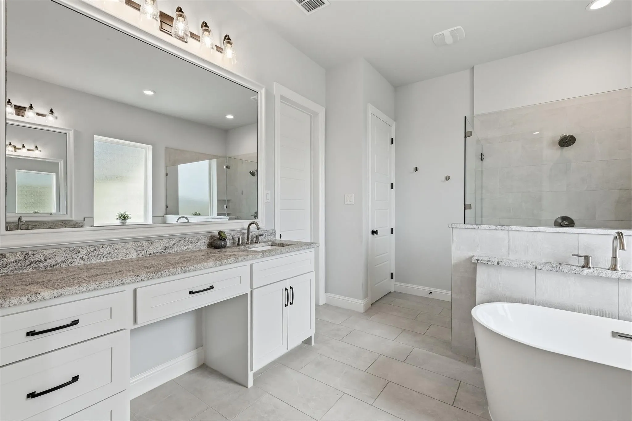 Spacious bathroom featuring a dual vanity with stone countertops, a large frameless mirror, a freestanding soaking tub, a separate walk-in shower with a glass enclosure, and light-toned tile flooring