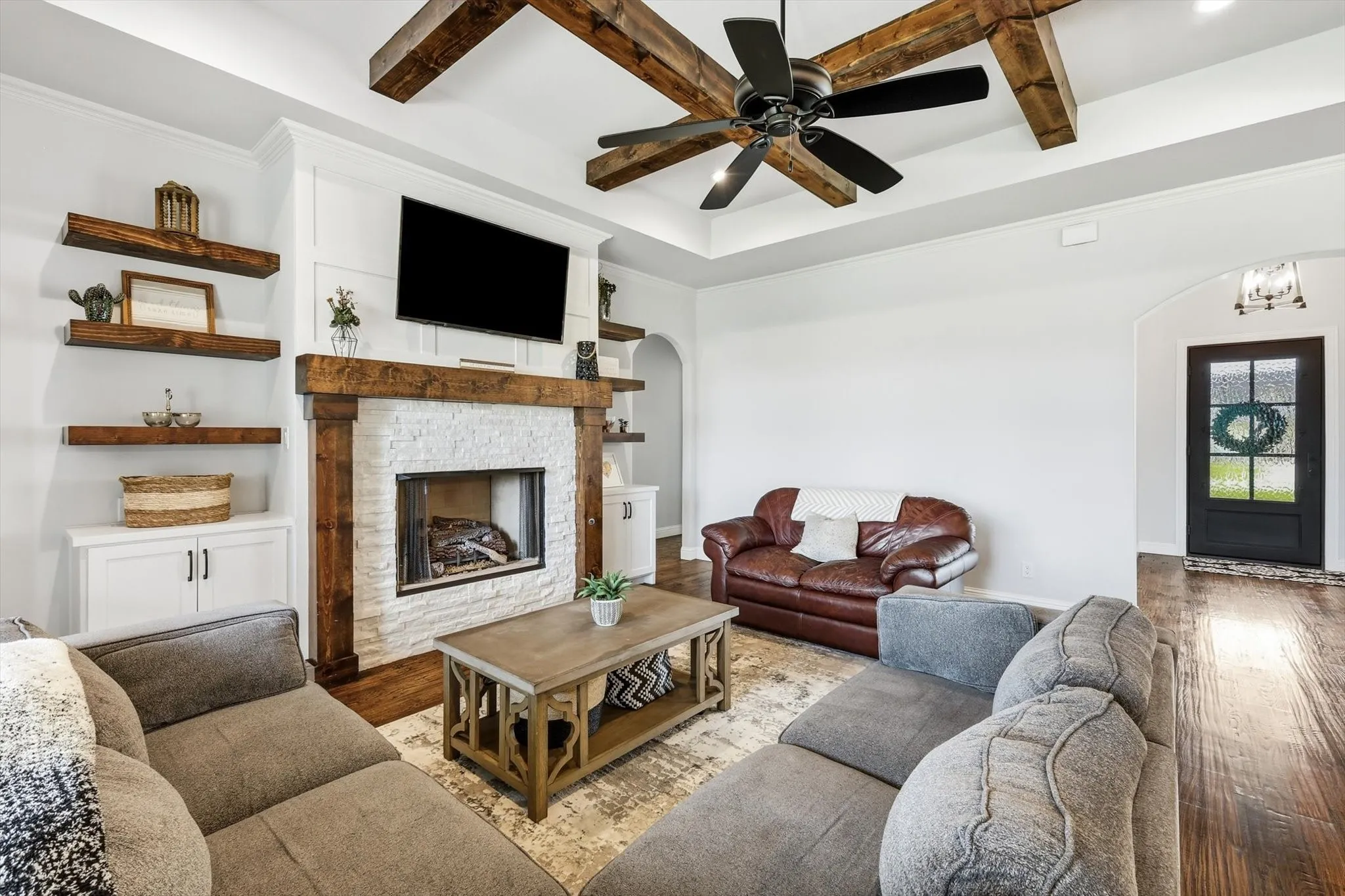 Living space featuring a whitewashed brick fireplace with a wood mantel, built-in shelving, wood-finish flooring, and a tray ceiling with exposed wood beams and a ceiling fan