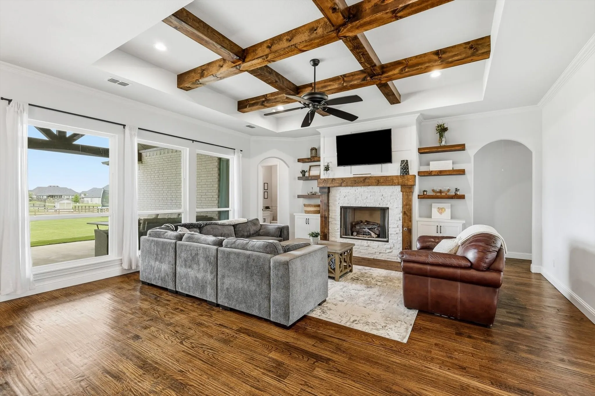Spacious living area featuring exposed wood ceiling beams, a white brick fireplace with a wood mantel, built-in shelving, wood-finish flooring, and large windows