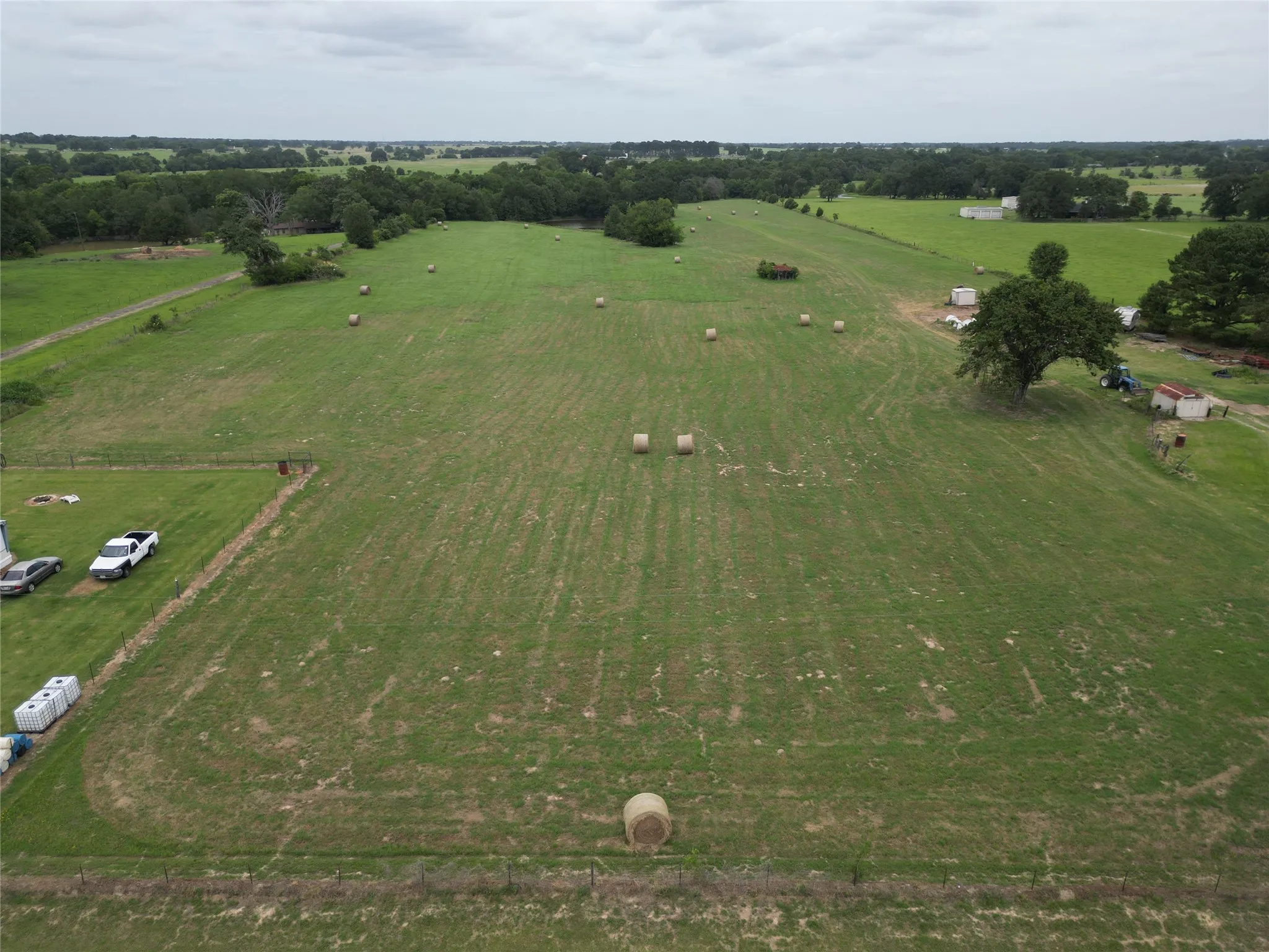 Expansive green acreage featuring a perimeter fence, scattered mature trees, and visible baled hay
