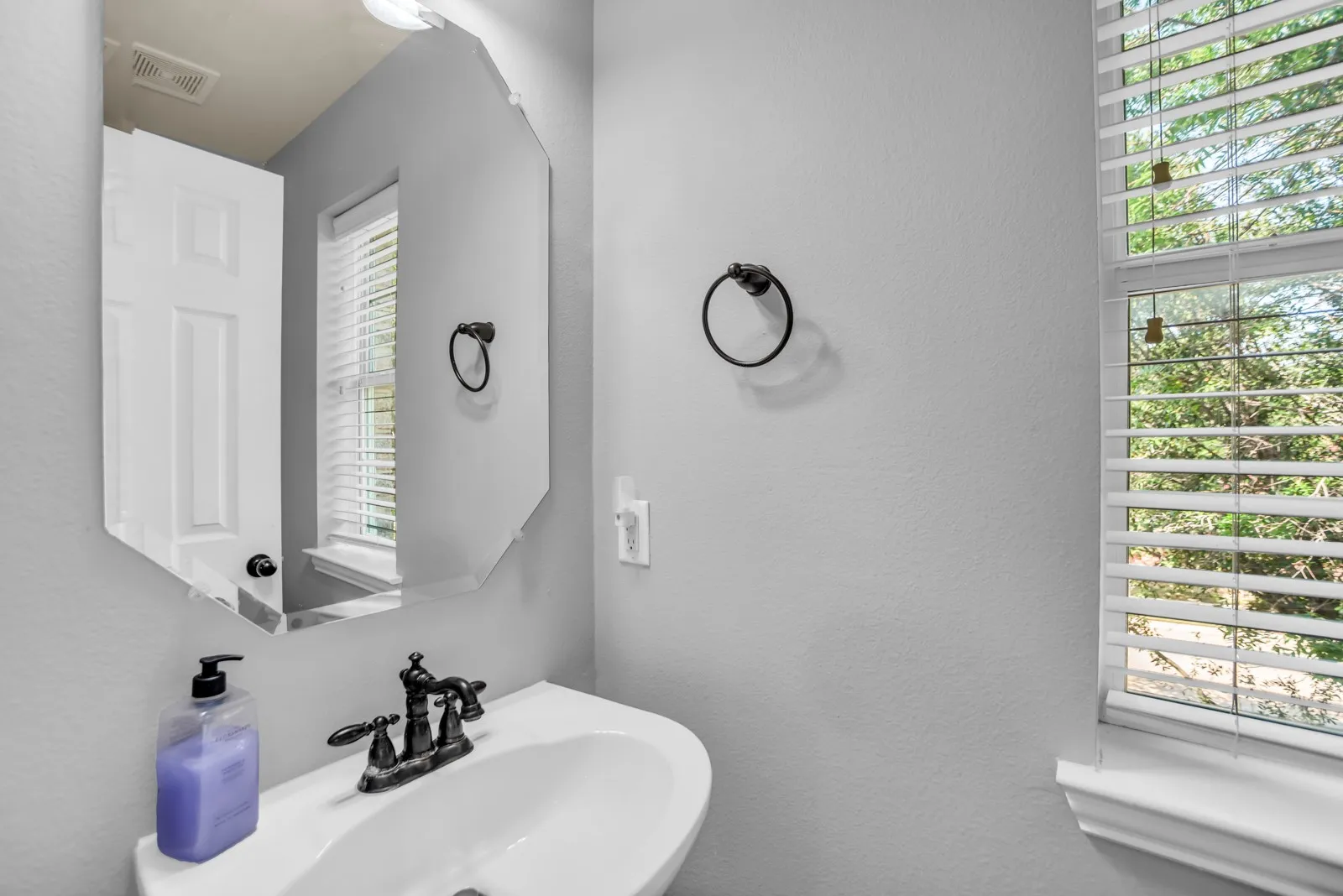 Pedestal sink featuring a dark-finish faucet, an octagonal frameless mirror, and a window with horizontal blinds