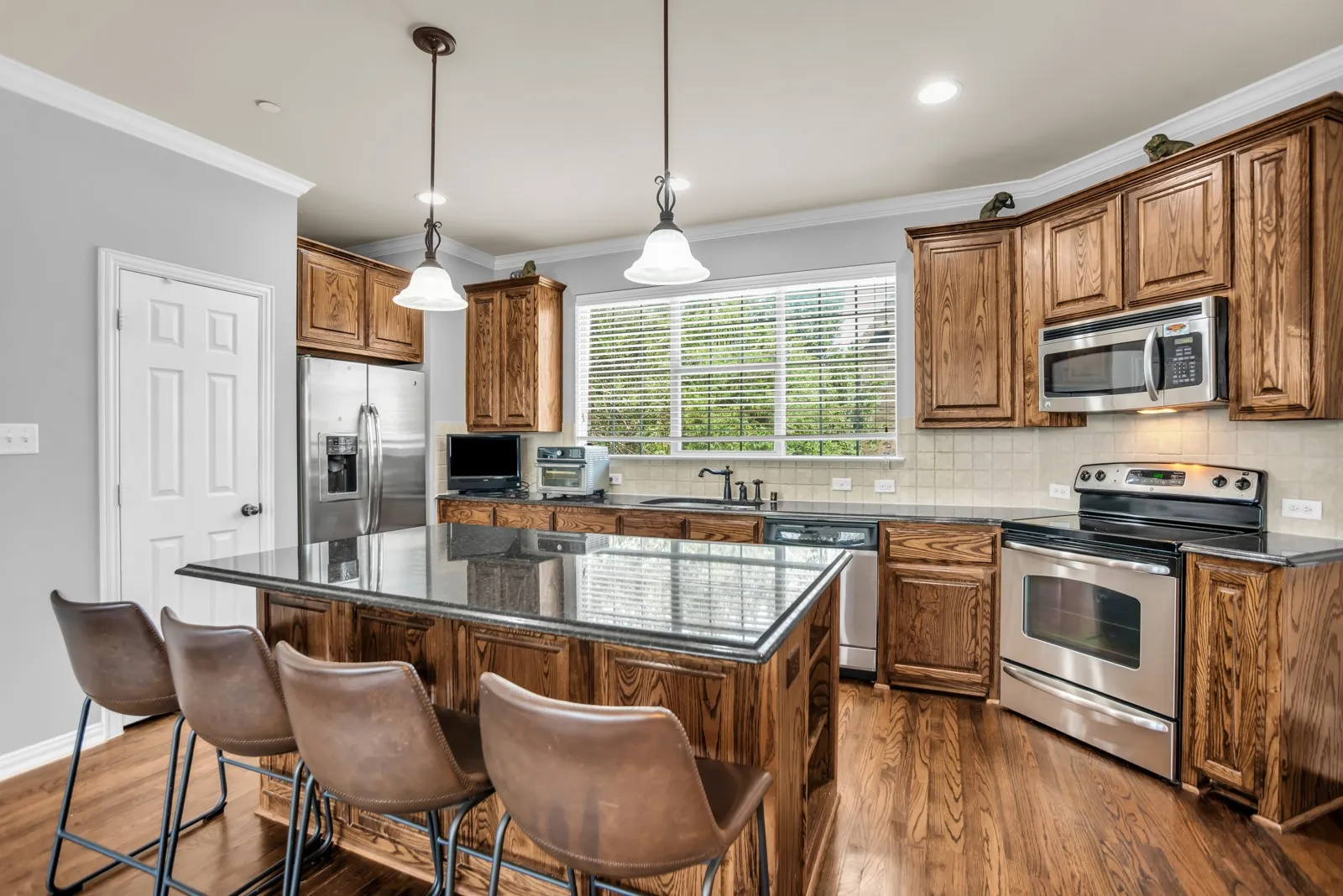 Kitchen featuring a substantial island with a dark countertop, rich wood cabinetry, stainless steel appliances, wood-finish flooring, and a window with horizontal blinds