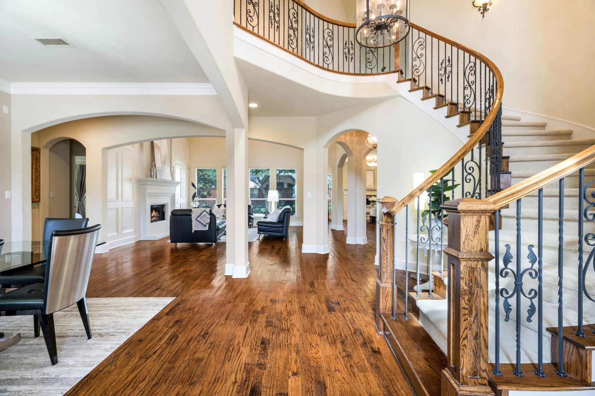 Curved staircase with wrought iron balusters and wood handrails, wood-finish flooring, arched doorways, a fireplace with a white mantel, and ceiling-mounted chandeliers