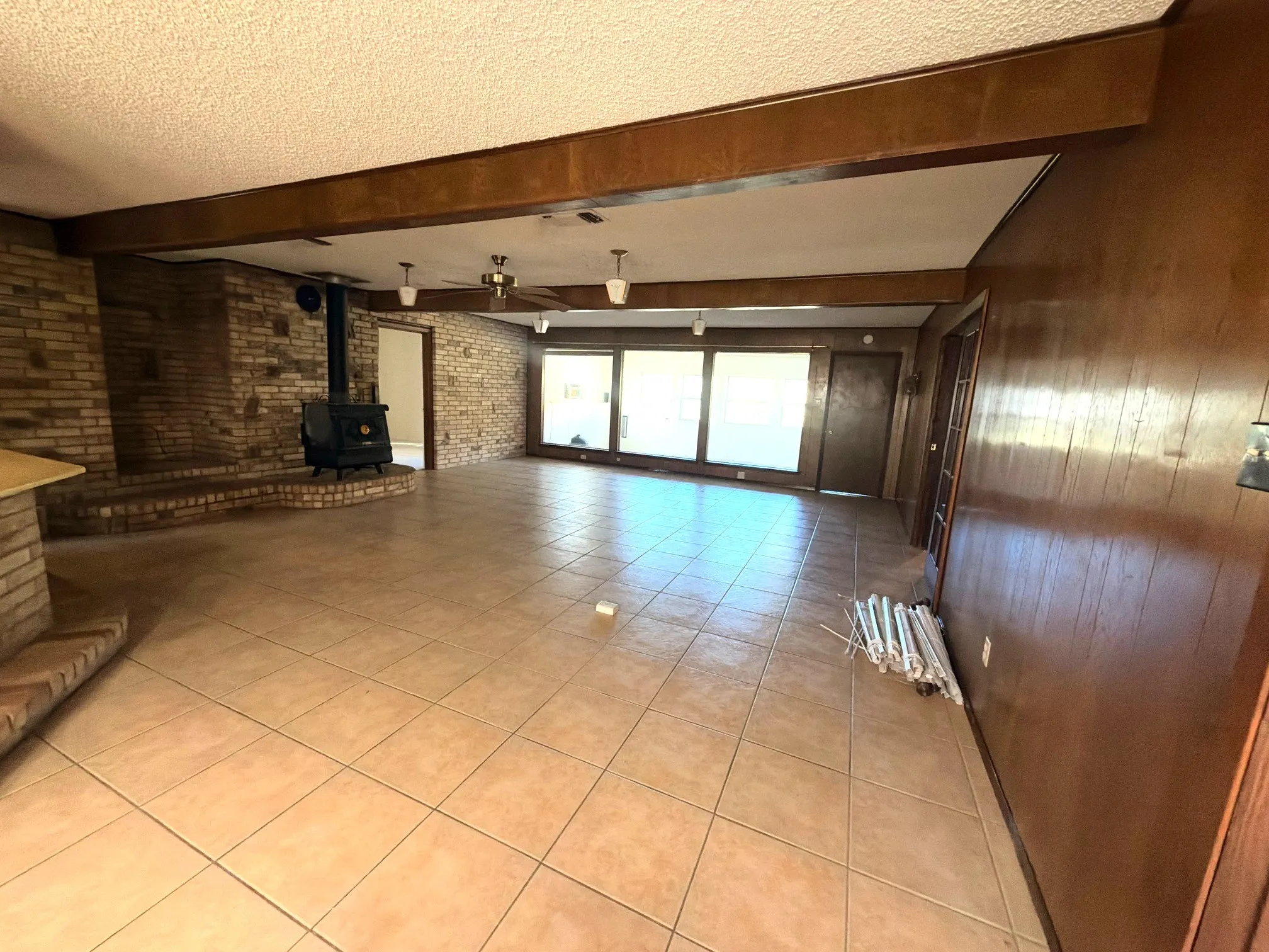 Brick accent wall with hearth, tile flooring, wood-paneled walls, exposed wood beams, and a wood-burning stove