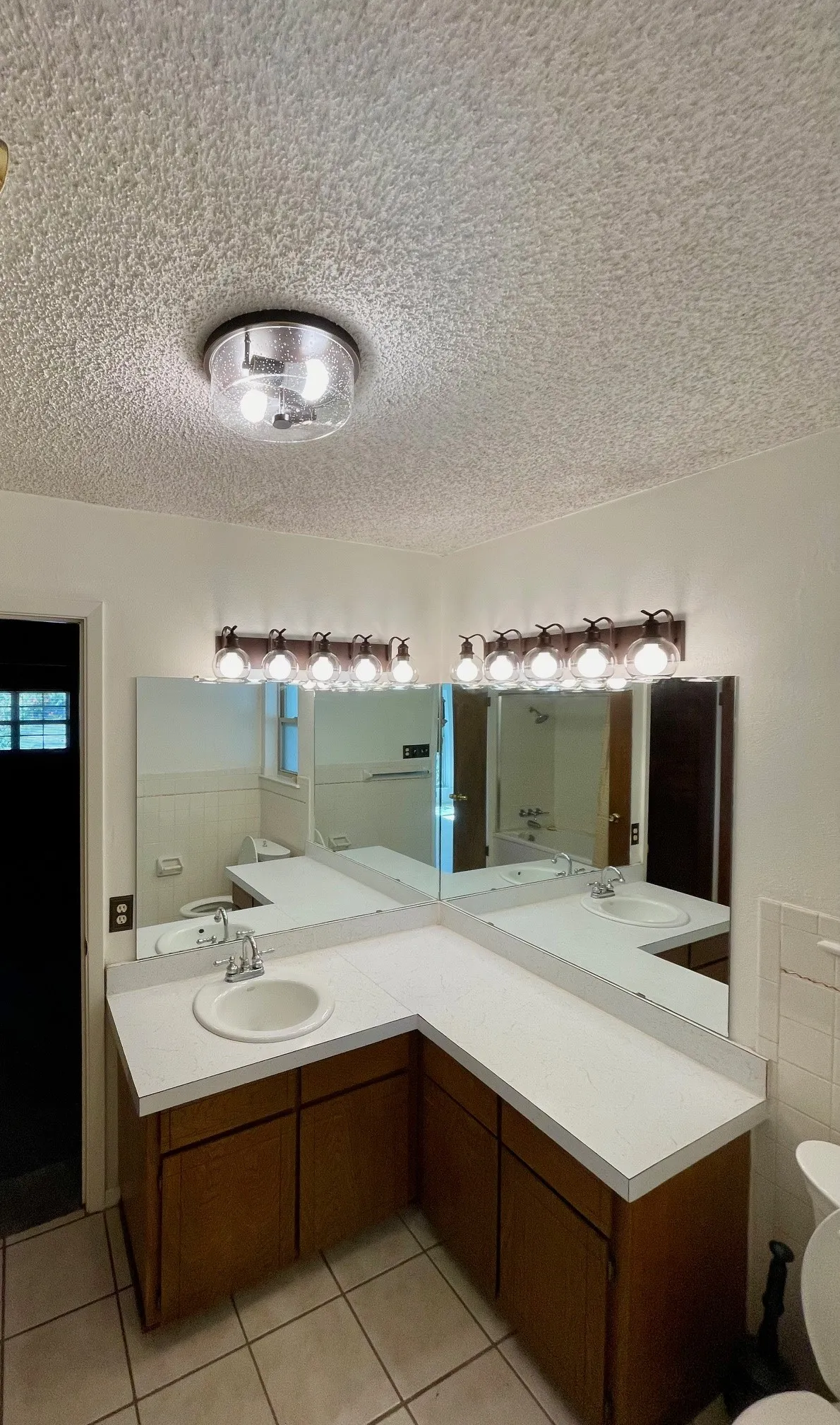 Dual vanity with white countertop and integrated sinks, wood-finish cabinetry, and two multi-bulb light fixtures