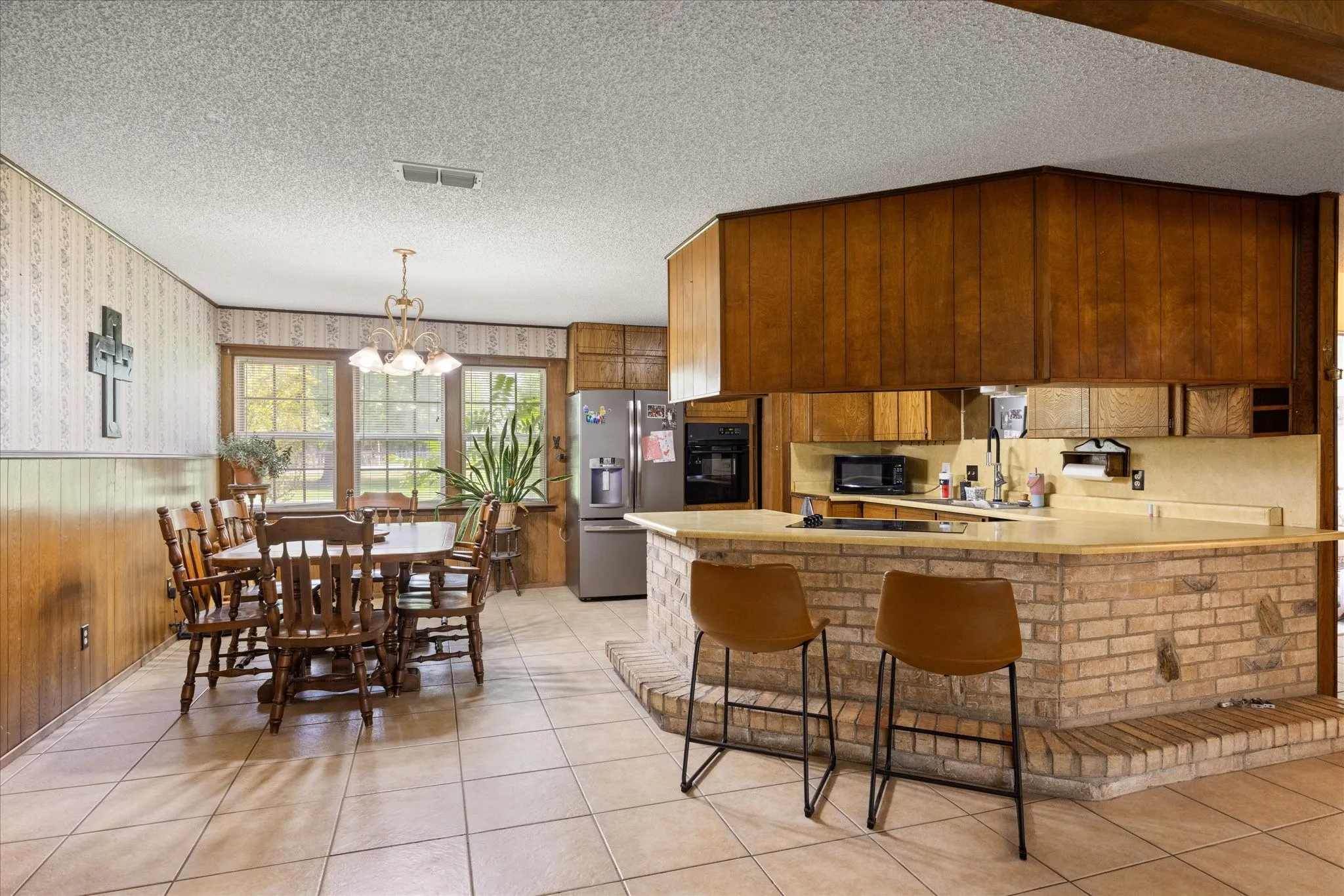 Spacious kitchen and dining area featuring a brick island with countertop seating, wood-finish cabinetry, tile flooring, and multiple windows