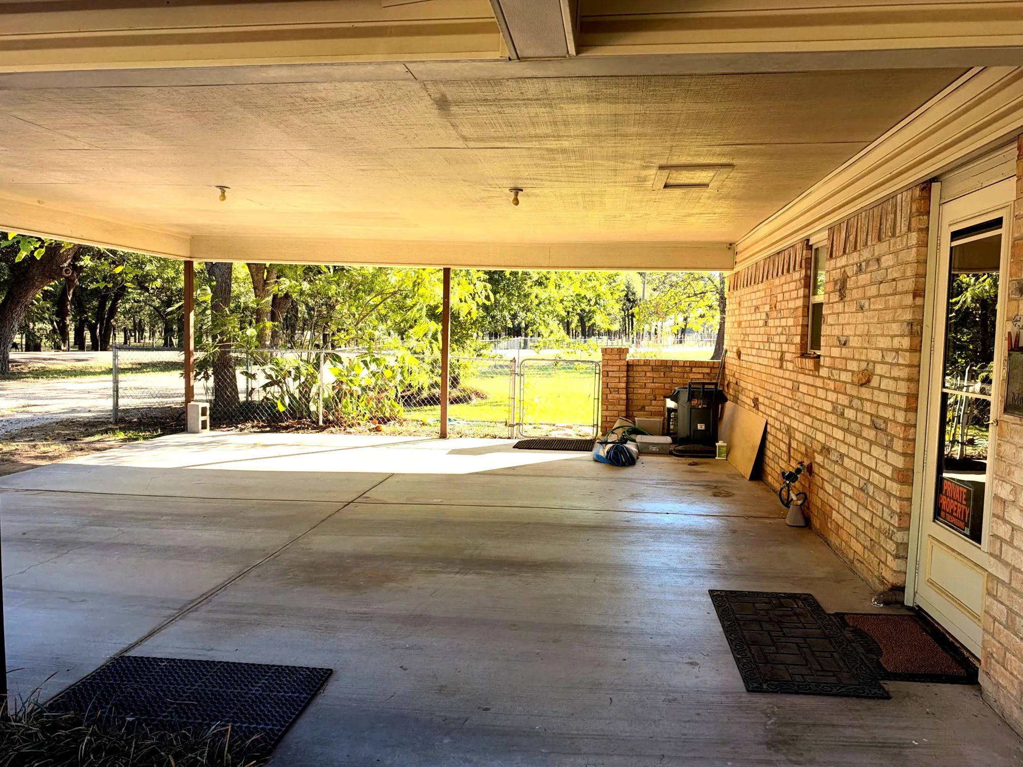 Covered concrete patio with a brick exterior, framed by mature trees, and featuring a wrought iron fence