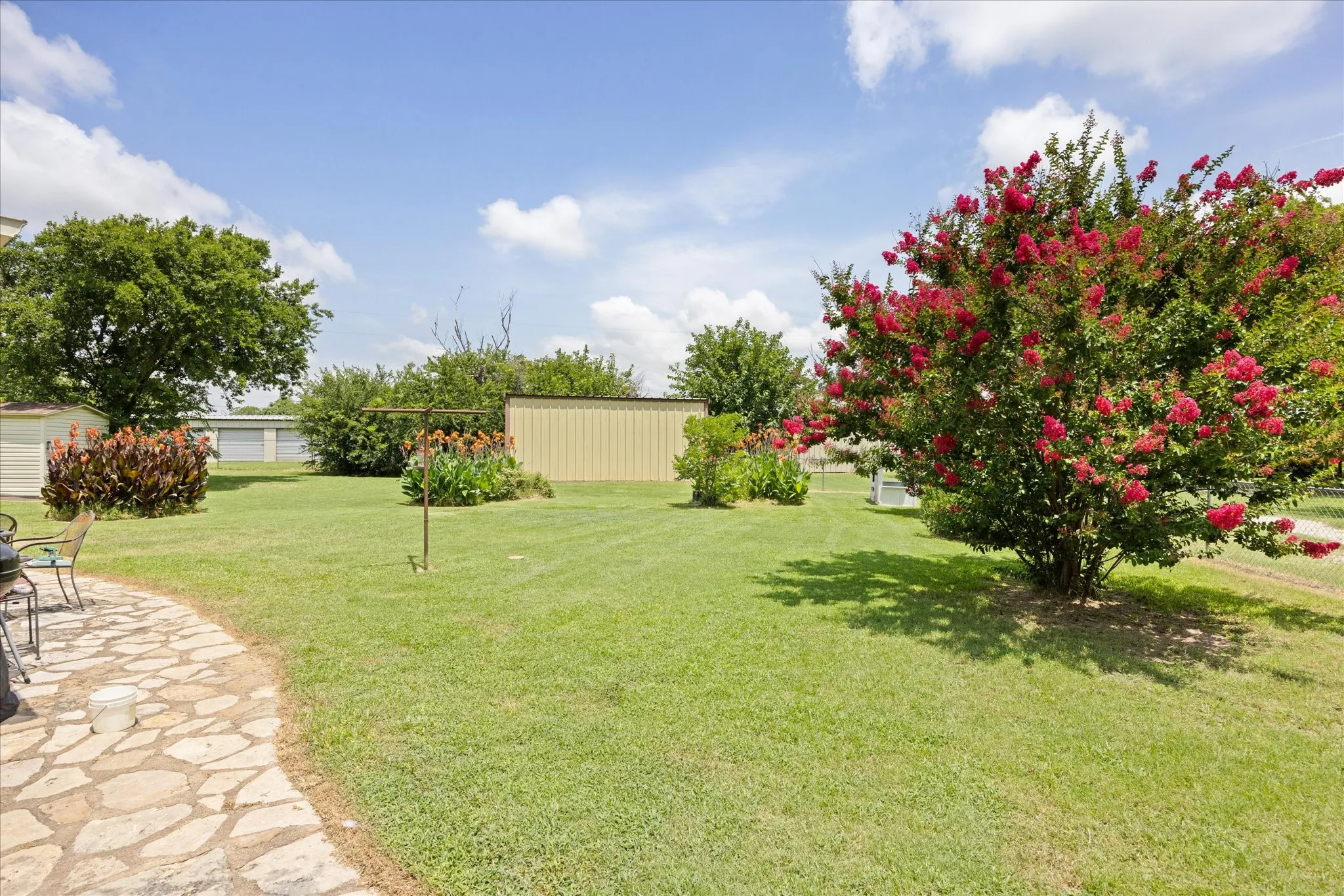 Expansive grassy yard featuring a stone patio, mature trees, flowering shrubs, and two storage sheds