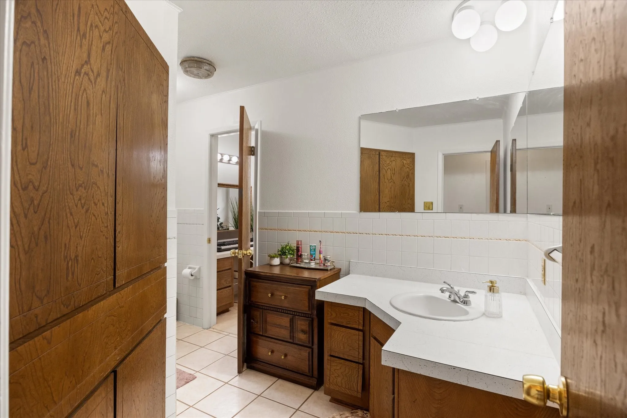 Bathroom featuring a white countertop with integrated sink, wood-finish cabinetry, a large wall-mounted mirror, white square tile wainscoting, and a built-in wood-finish linen cabinet