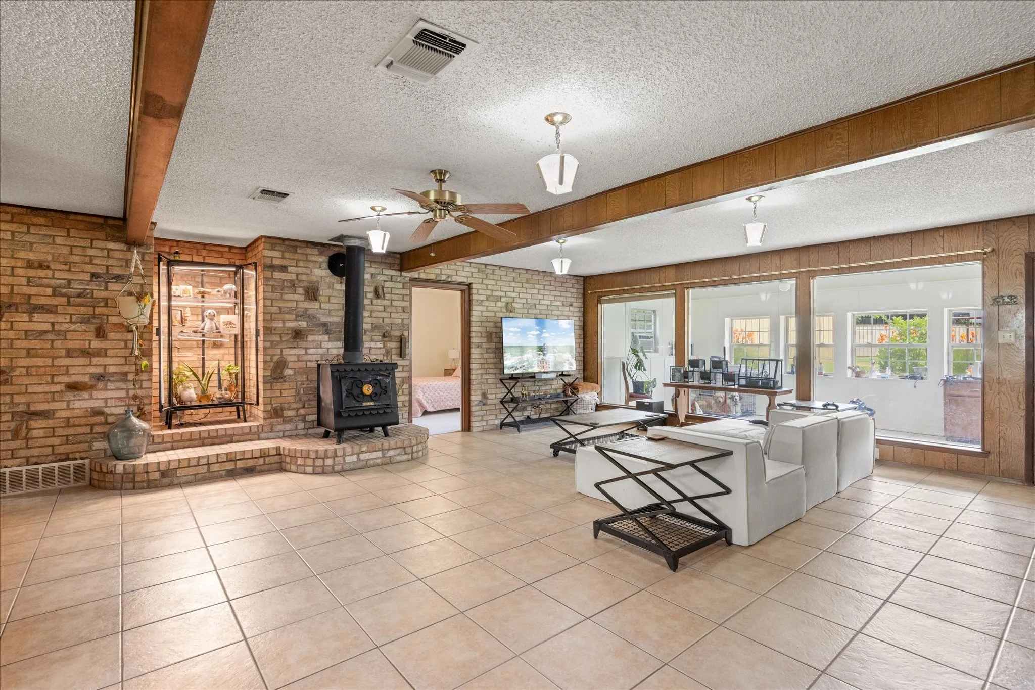 Spacious living area featuring light-toned tile flooring, exposed wood beams, and a brick accent wall with an integrated wood-burning stove on a raised hearth