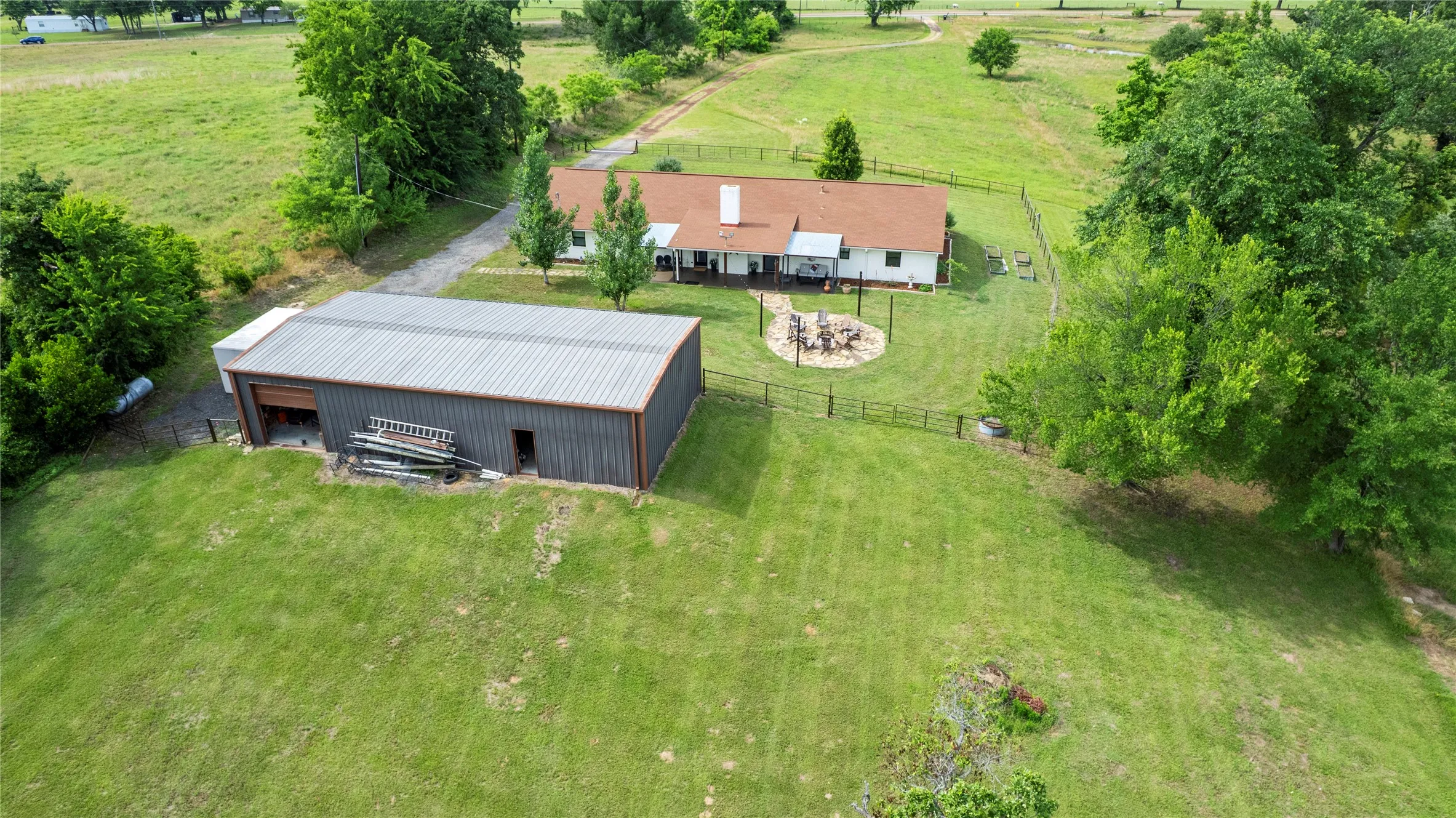Expansive green landscape featuring a detached metal-sided workshop with a roll-up door, a single-story residence with a red-shingled roof, a fenced backyard, and a gravel driveway