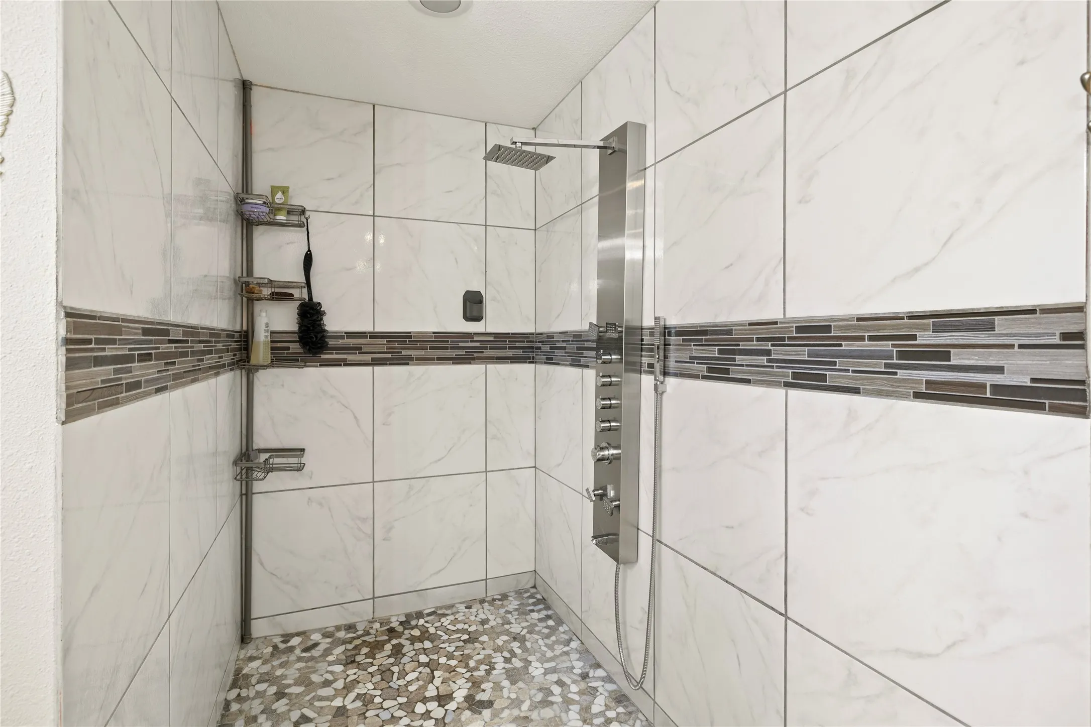 Walk-in shower featuring large format white tile walls with a mosaic accent band, a pebble-finish floor, and a multi-function shower panel with a rainfall head