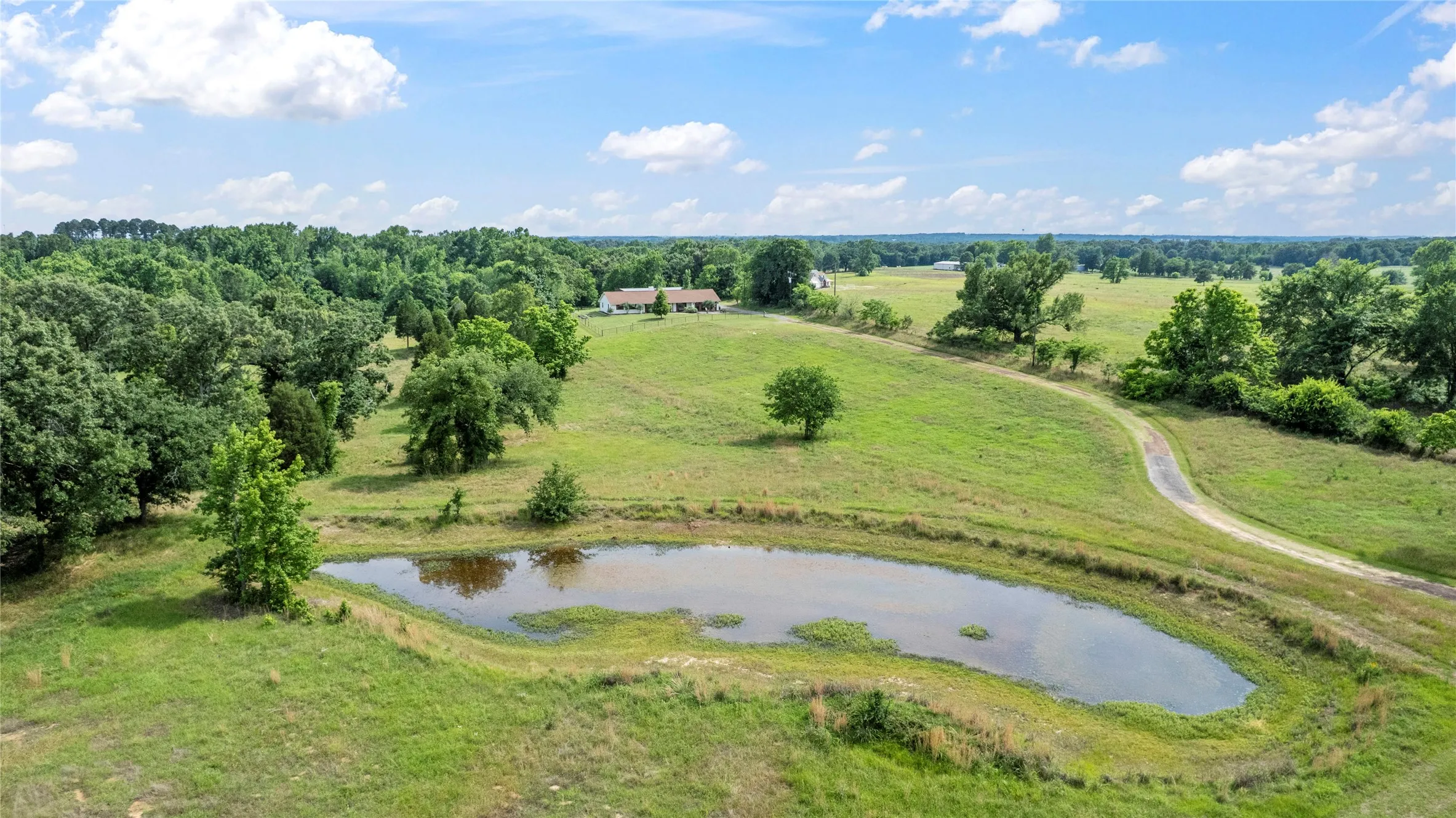 Expansive rural landscape featuring a natural pond, a winding driveway, scattered mature trees, and a distant single-story residence
