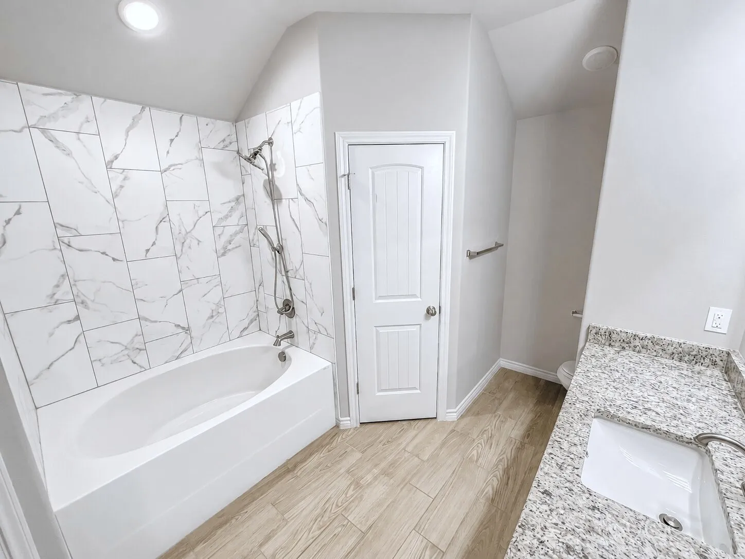 Bathroom featuring a built-in tub with a shower head, white marble-style wall tiling, a light wood-finish floor, a white vanity with an undermount sink, and a speckled countertop