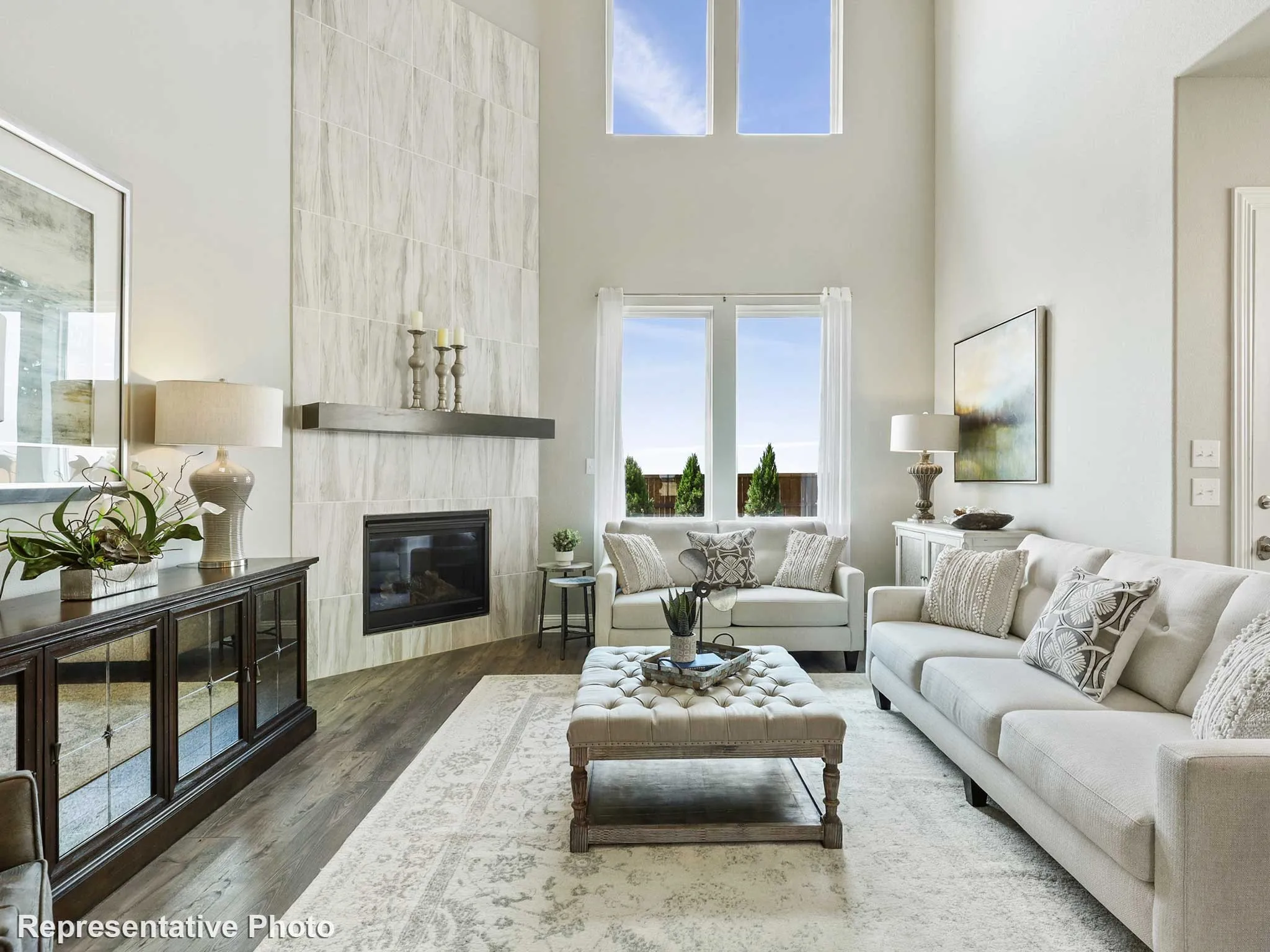 Spacious living area featuring a two-story ceiling, large windows, a tiled fireplace with a dark wood mantel, and wood-finish flooring