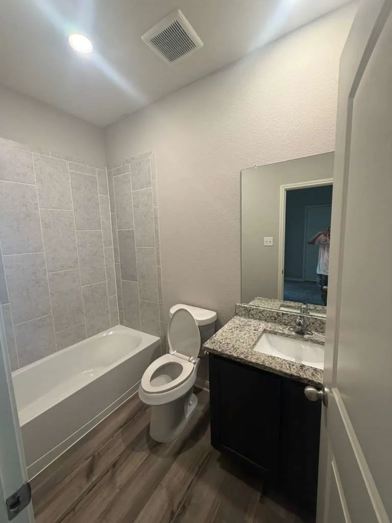 Bathroom featuring a white bathtub, light gray subway tile surround, wood-finish flooring, and a single vanity with a granite countertop and dark cabinetry
